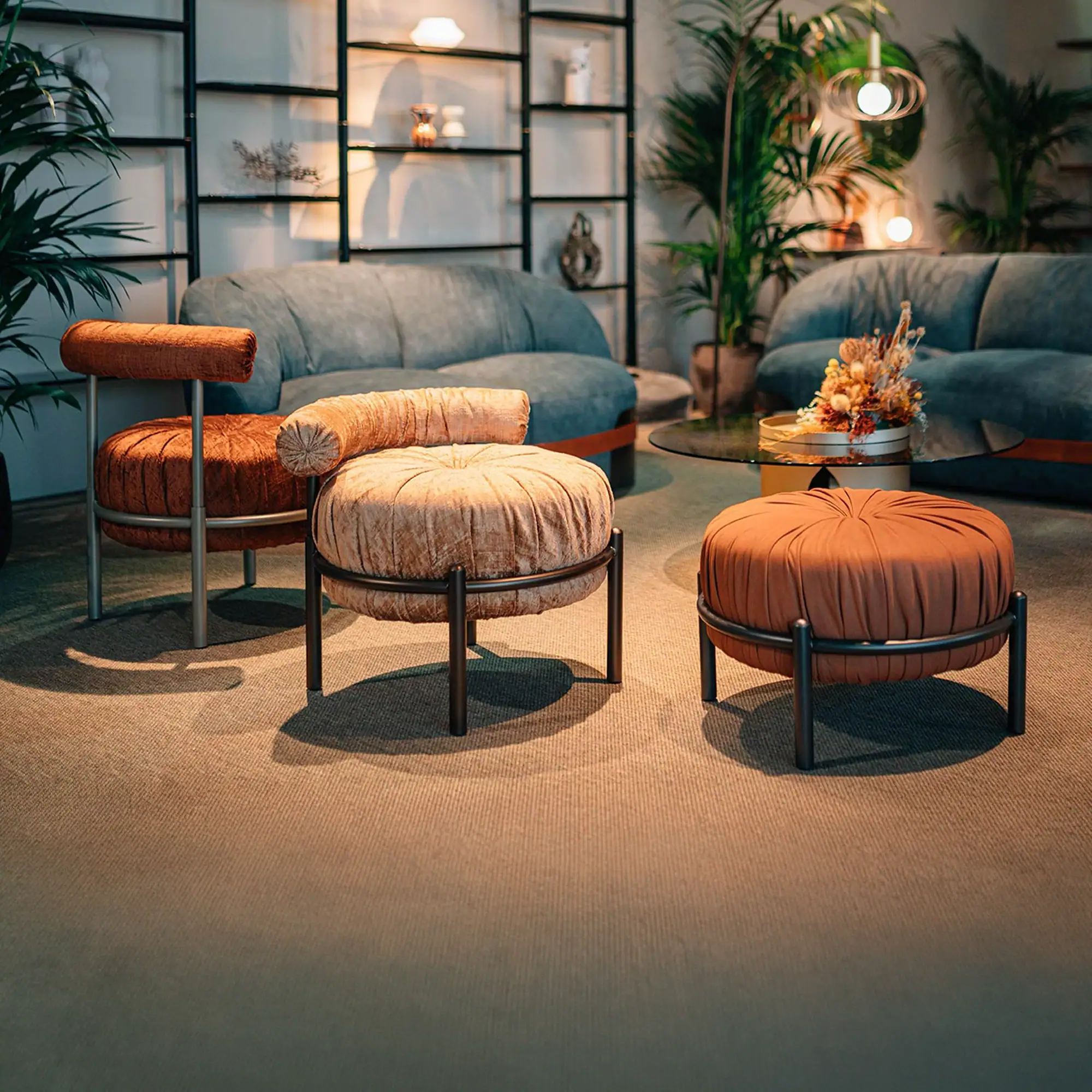 A living room with three round, upholstered stools in rust and peach colors, two curved blue sofas, a glass coffee table with dried flowers, and potted palm plants.
