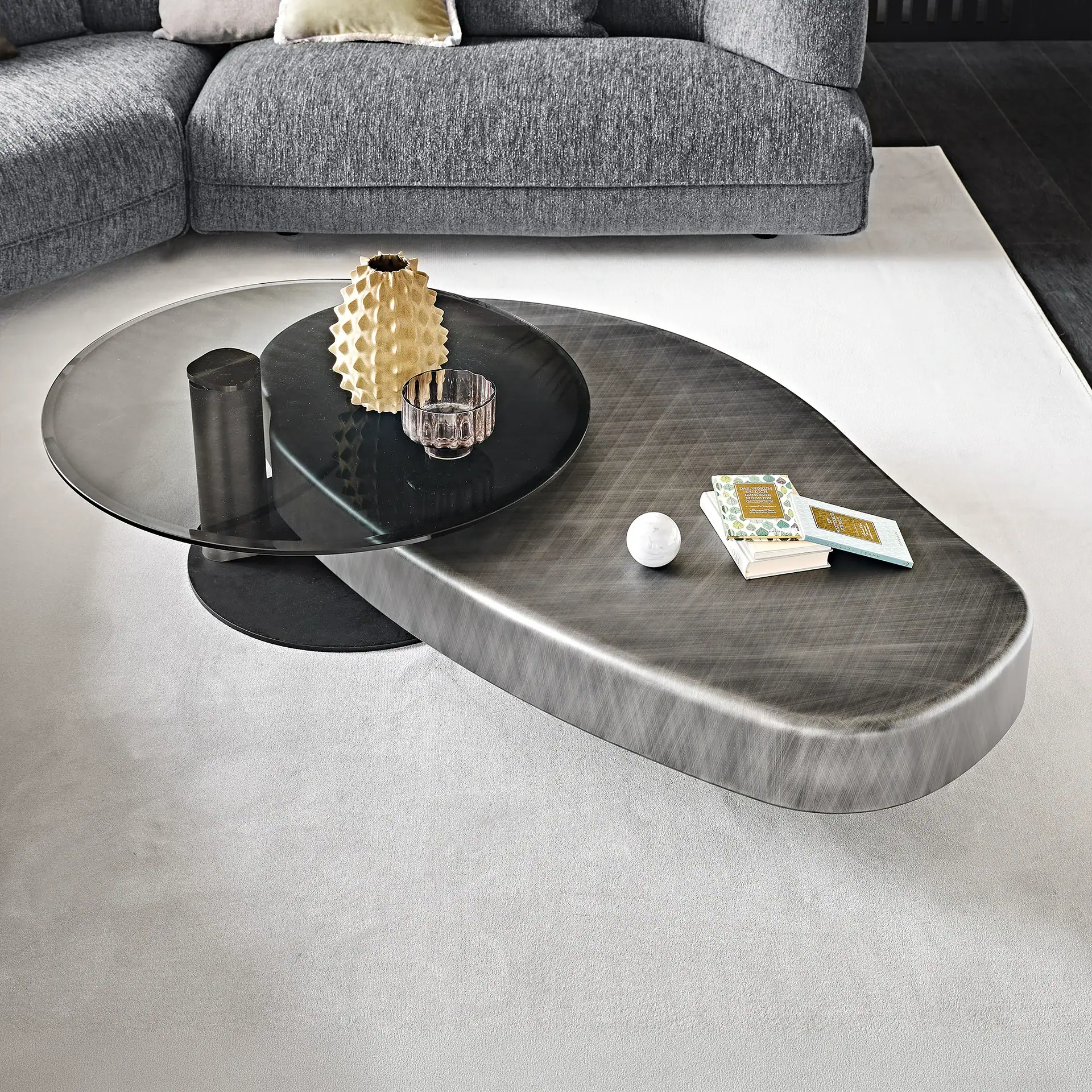 A modern, two-tiered coffee table with a dark metallic base and a smoked glass top, adorned with a gold vase, glass cup, marble sphere, and books, set on a light grey rug in front of a grey sofa.
