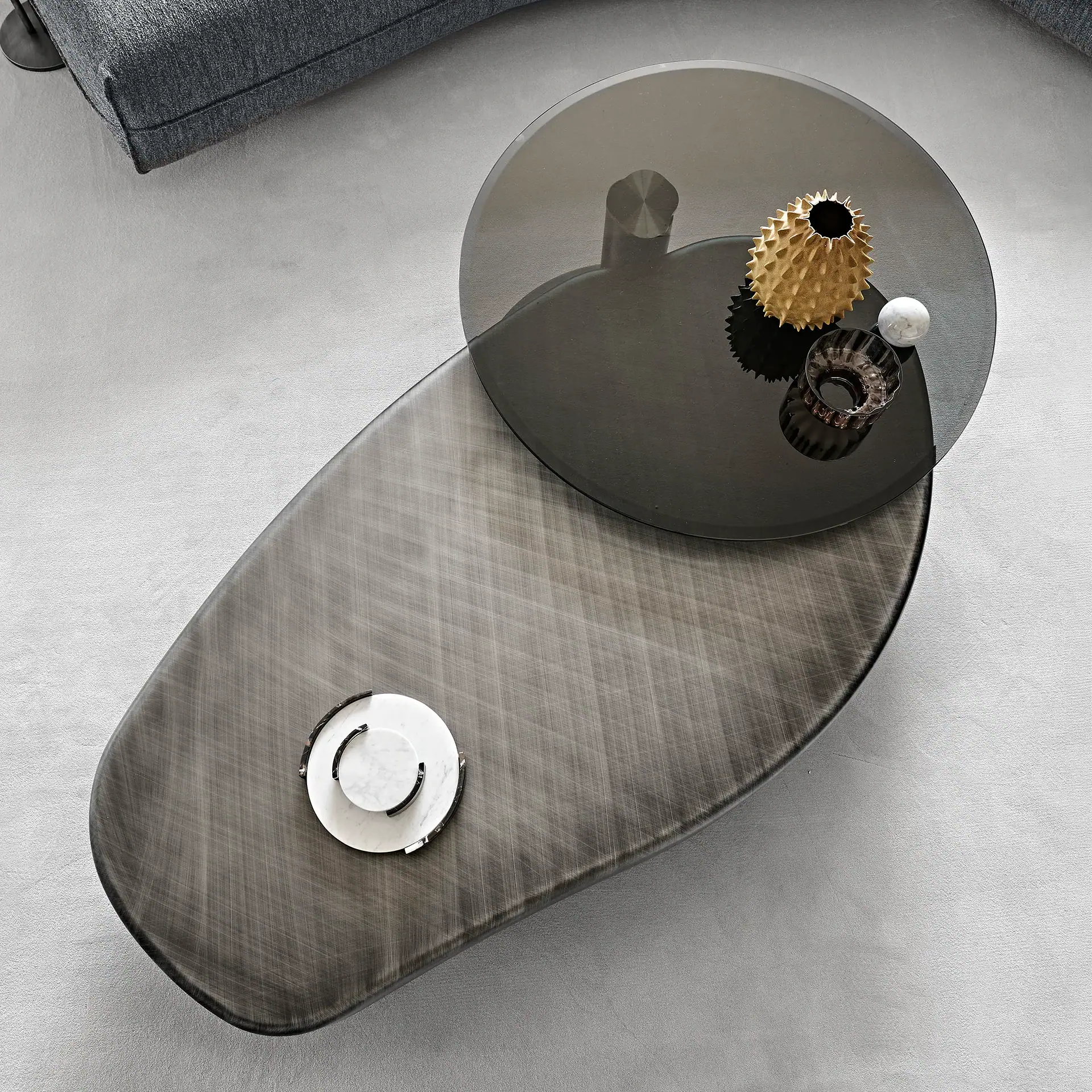 Overhead view of a modern, two-tiered dark grey glass coffee table. The lower table is elongated, and the upper is circular, holding a golden spiky vase, a marble sphere, and a glass votive. A grey sofa is partially visible.