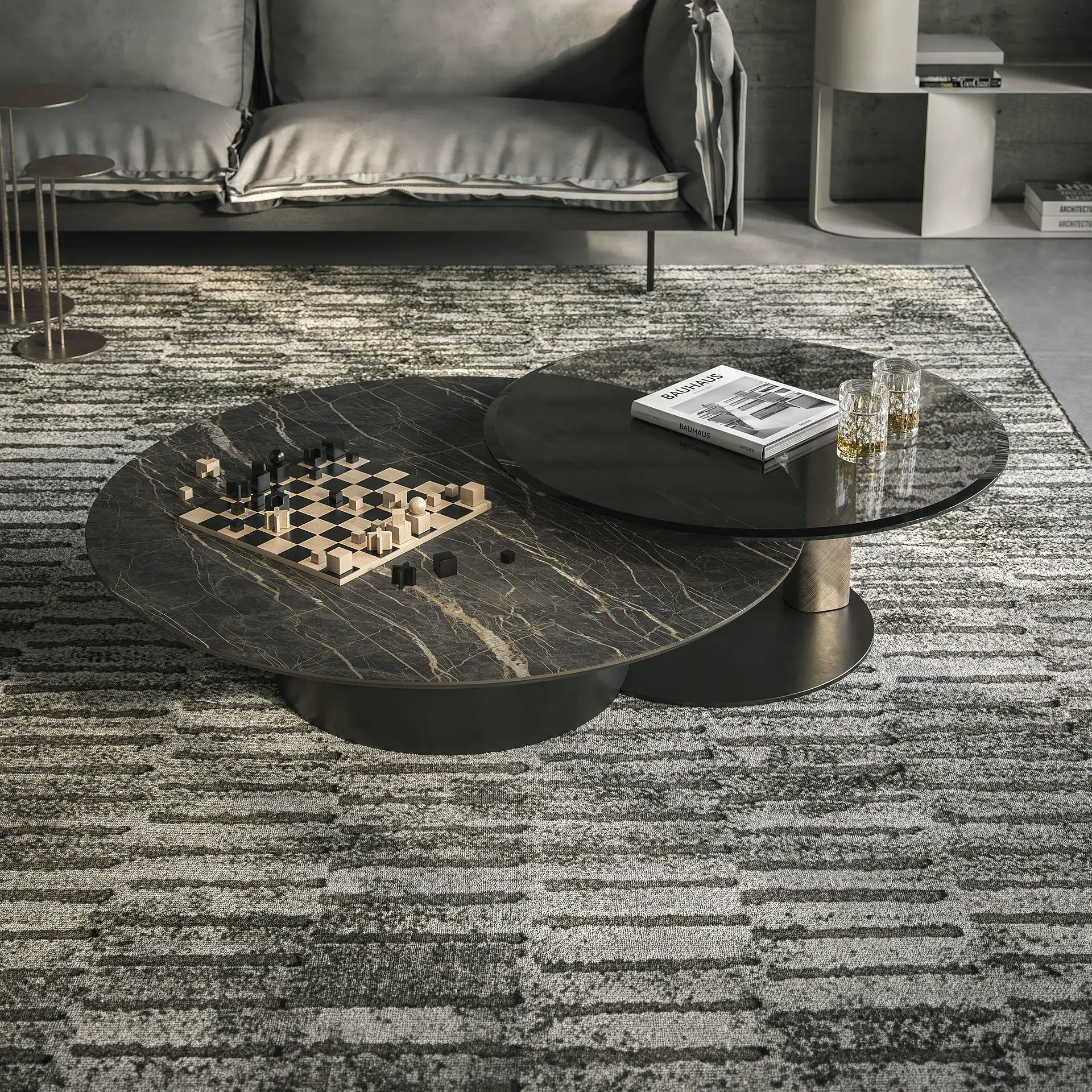 Two round, overlapping coffee tables in a modern living room. The lower table is dark marble with a wooden chess set, and the upper is dark glass with a 'Bauhaus' book and two drinks. A grey sofa and patterned rug complete the scene.