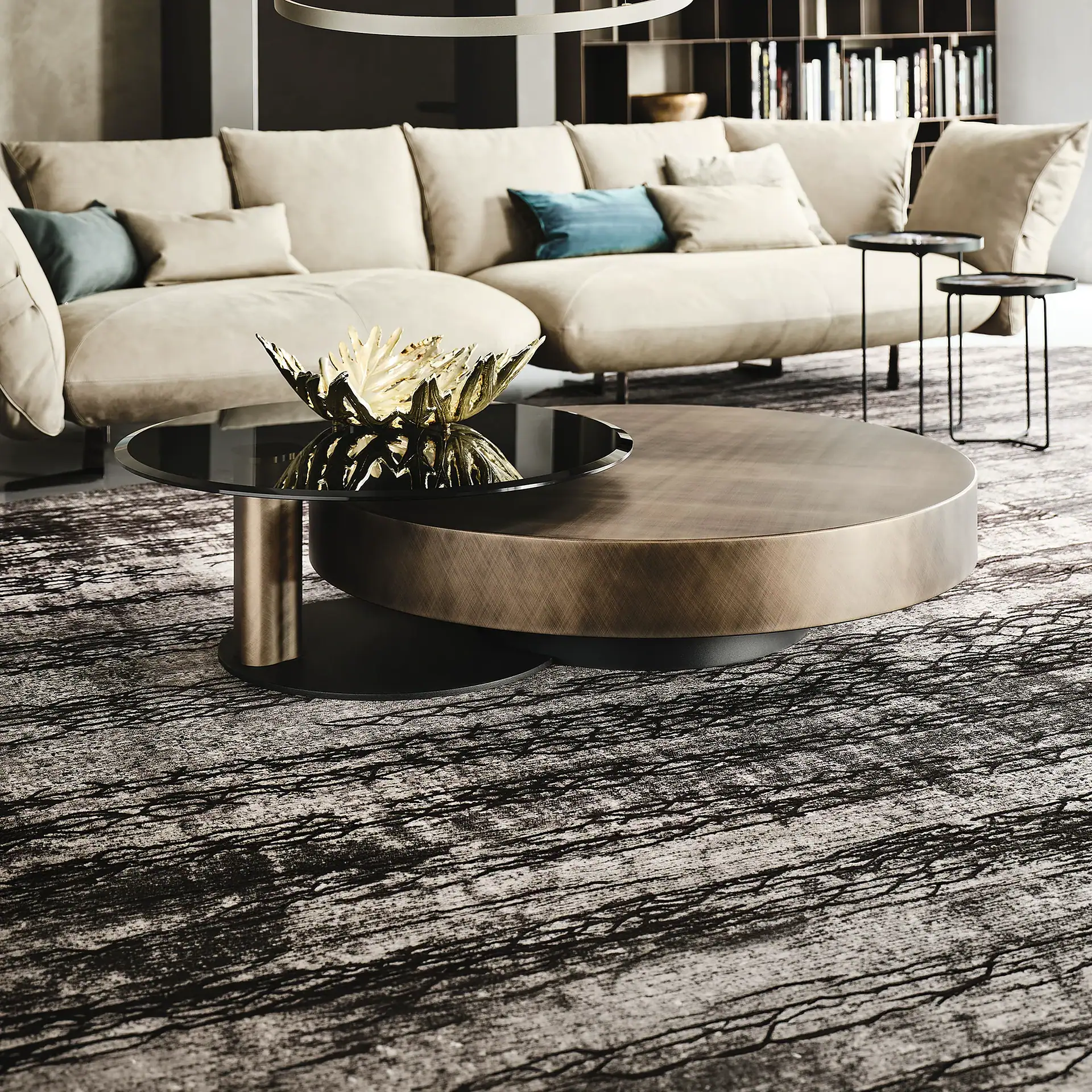 A modern living room with a light beige sectional sofa, a multi-tiered round brass and glass coffee table with a golden decorative bowl, and a textured gray rug. A bookshelf is visible in the background.
