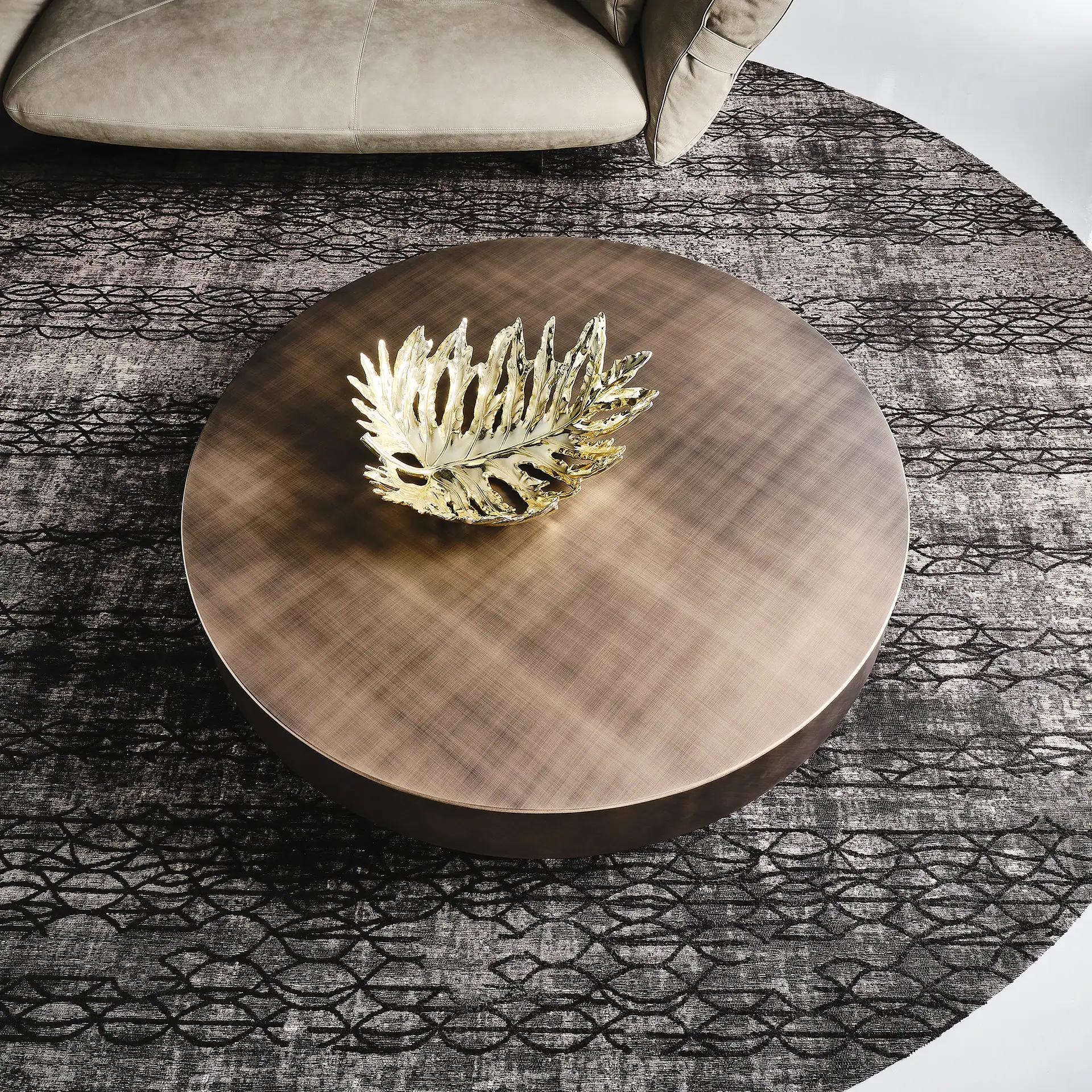 Overhead view of a round bronze coffee table with a golden leaf-shaped bowl, on a grey and black patterned rug, with a beige sofa visible.