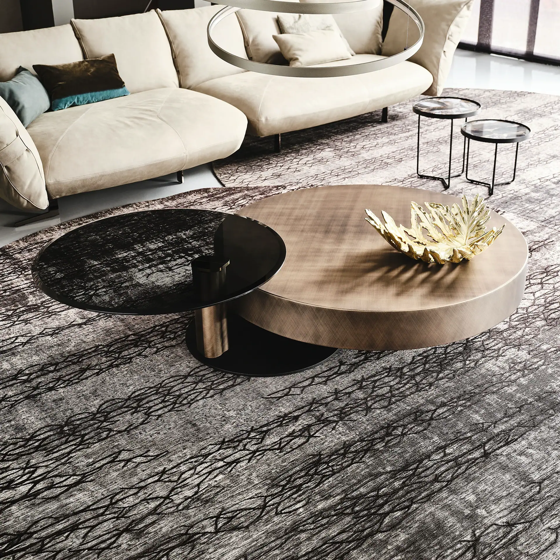 A modern living room with a cream curved sofa, overlapping circular coffee tables in bronze and black glass, and an abstract patterned rug.