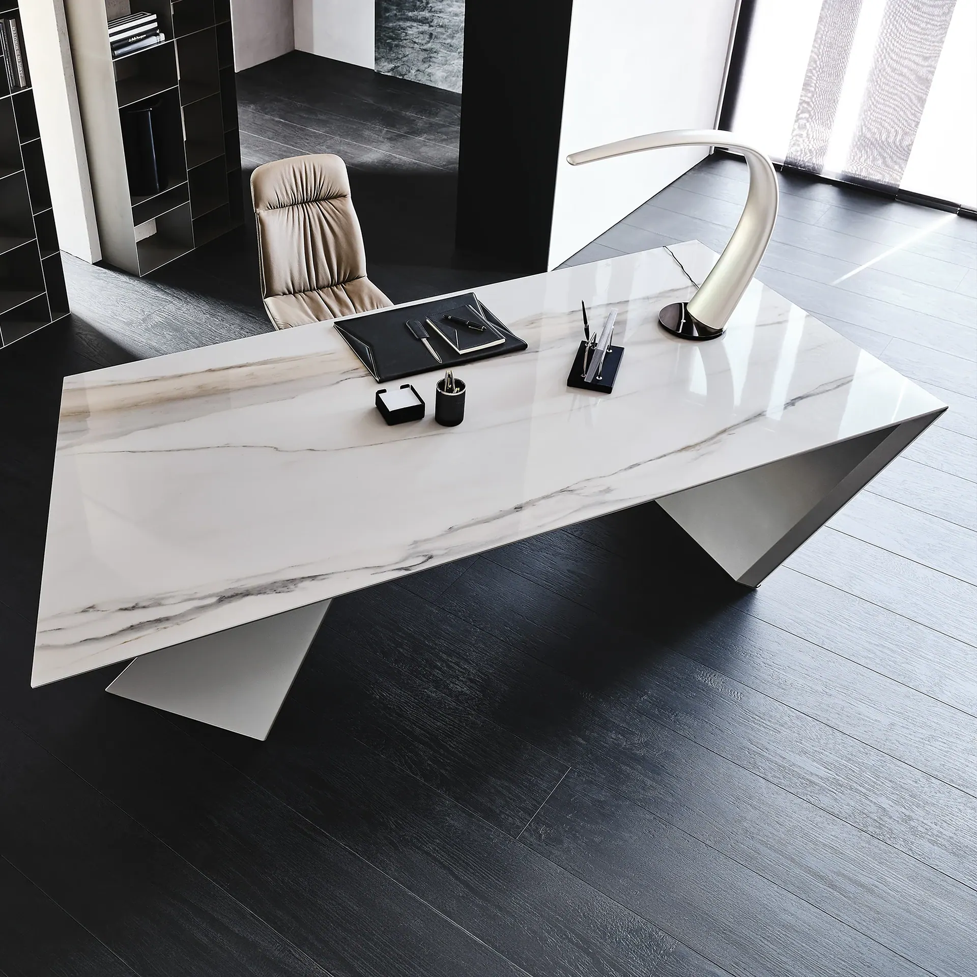 A modern office space featuring a large white marble-patterned desk with geometric legs, a tan leather office chair, and dark wood flooring. The desk holds office supplies and a silver lamp, with a shelving unit and window in the background.