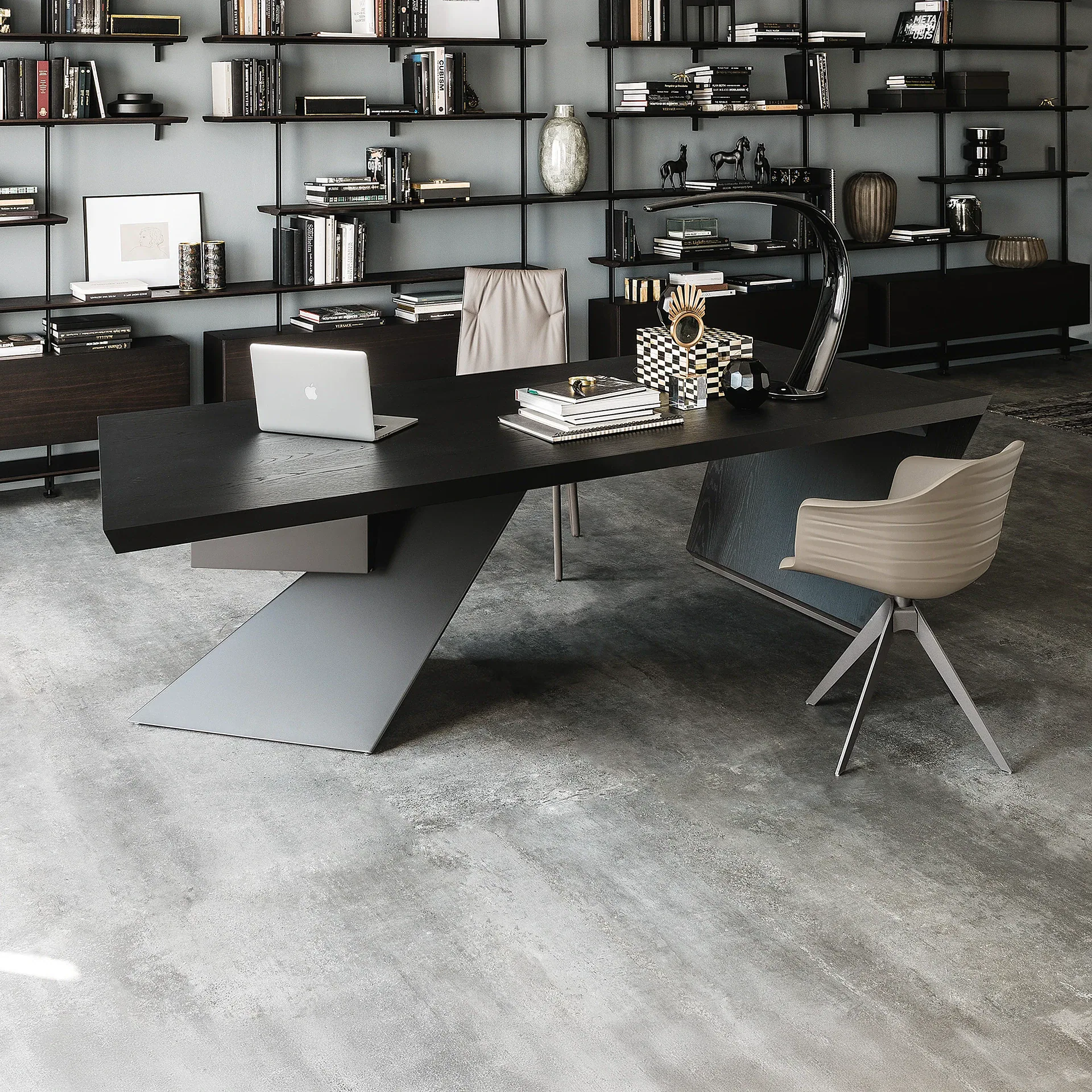 A modern home office with a large black wooden desk, a silver laptop, and a gold desk lamp. Two beige chairs are present, and the background features a light gray wall with black metal shelves filled with books and decorative items.