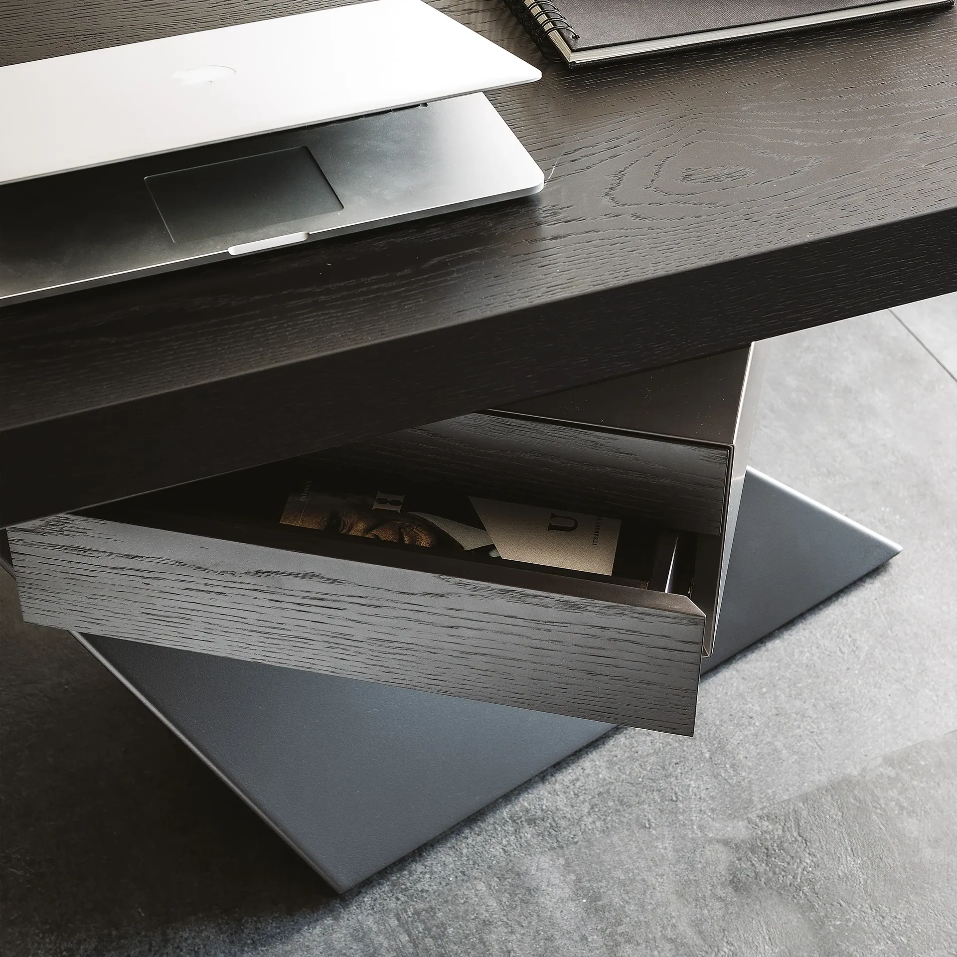A dark wood desk with a silver laptop, a black notebook, and a partially open drawer revealing items inside. The desk has modern metallic legs and sits on a gray tiled floor.
