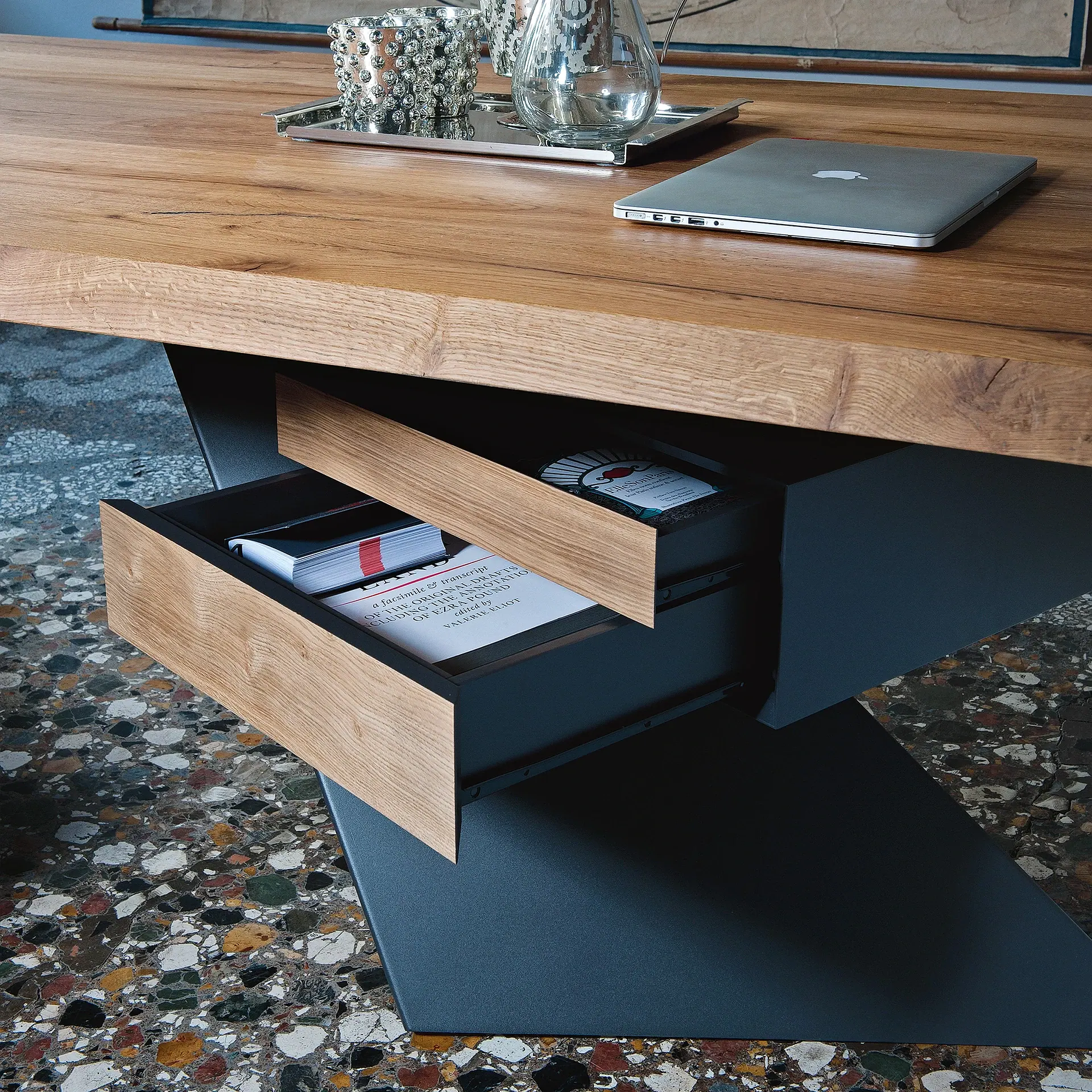 A wooden table with integrated drawers, a silver laptop, and decorative items on top, set against a colorful terrazzo floor.