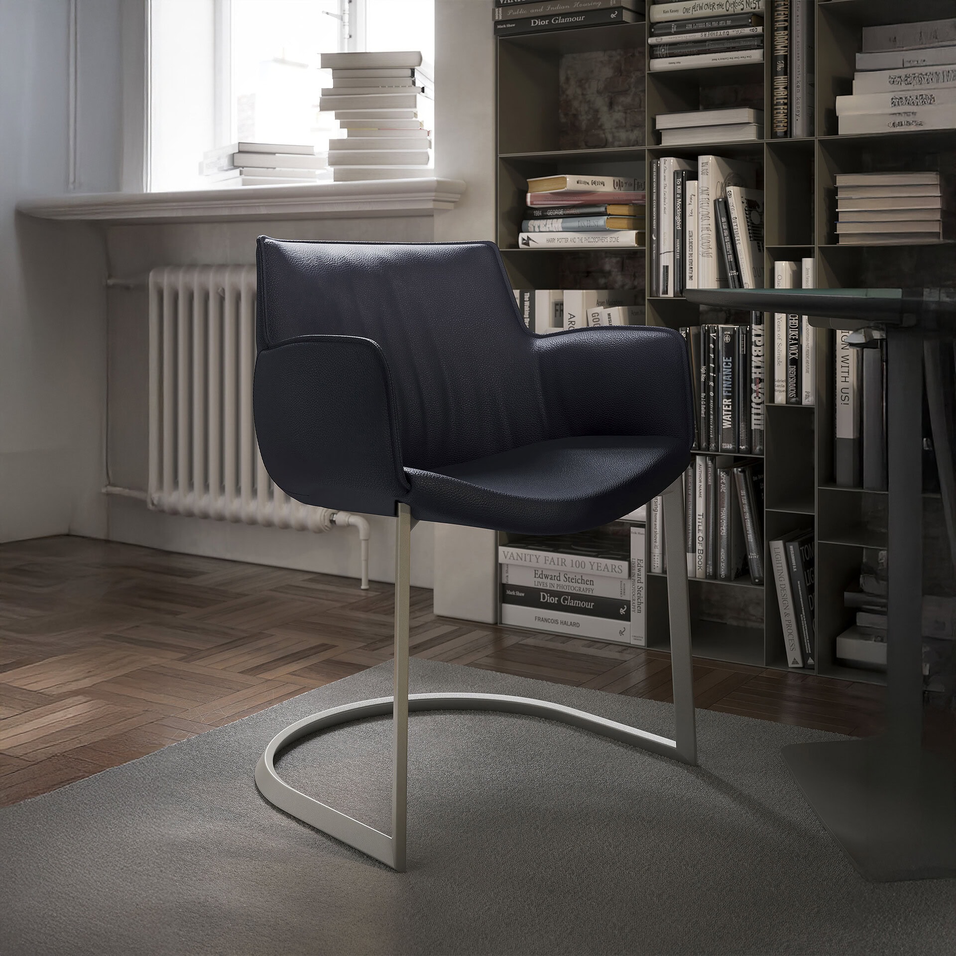A dark blue leather armchair with a cantilevered metal base sits on a gray rug in a bright room. A tall bookshelf filled with books is on the right, and a window with stacks of books on the sill is on the left.