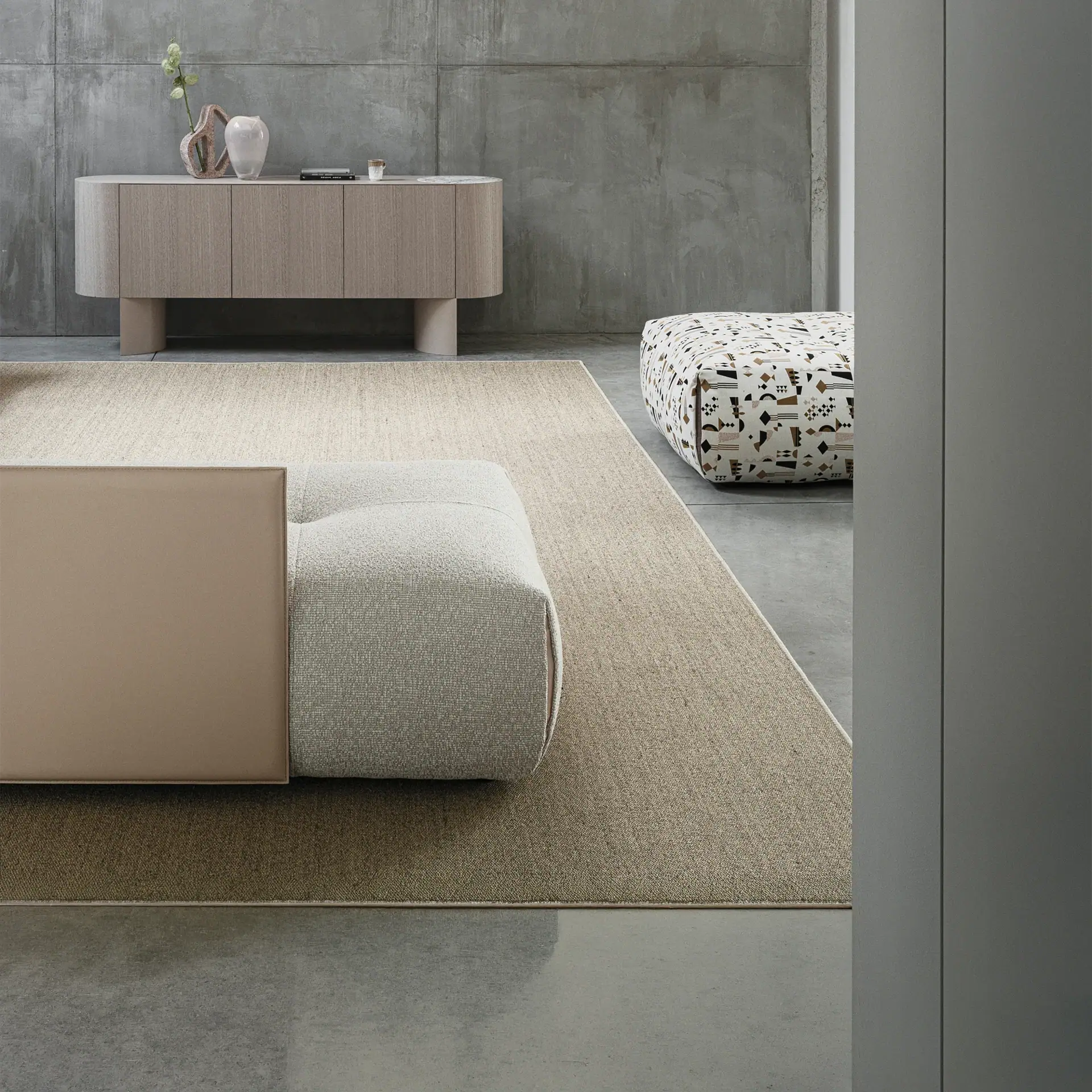 A modern living room with a light wooden sideboard, a textured beige rug, and modular seating. Concrete walls and a patterned ottoman complete the minimalist, neutral aesthetic.