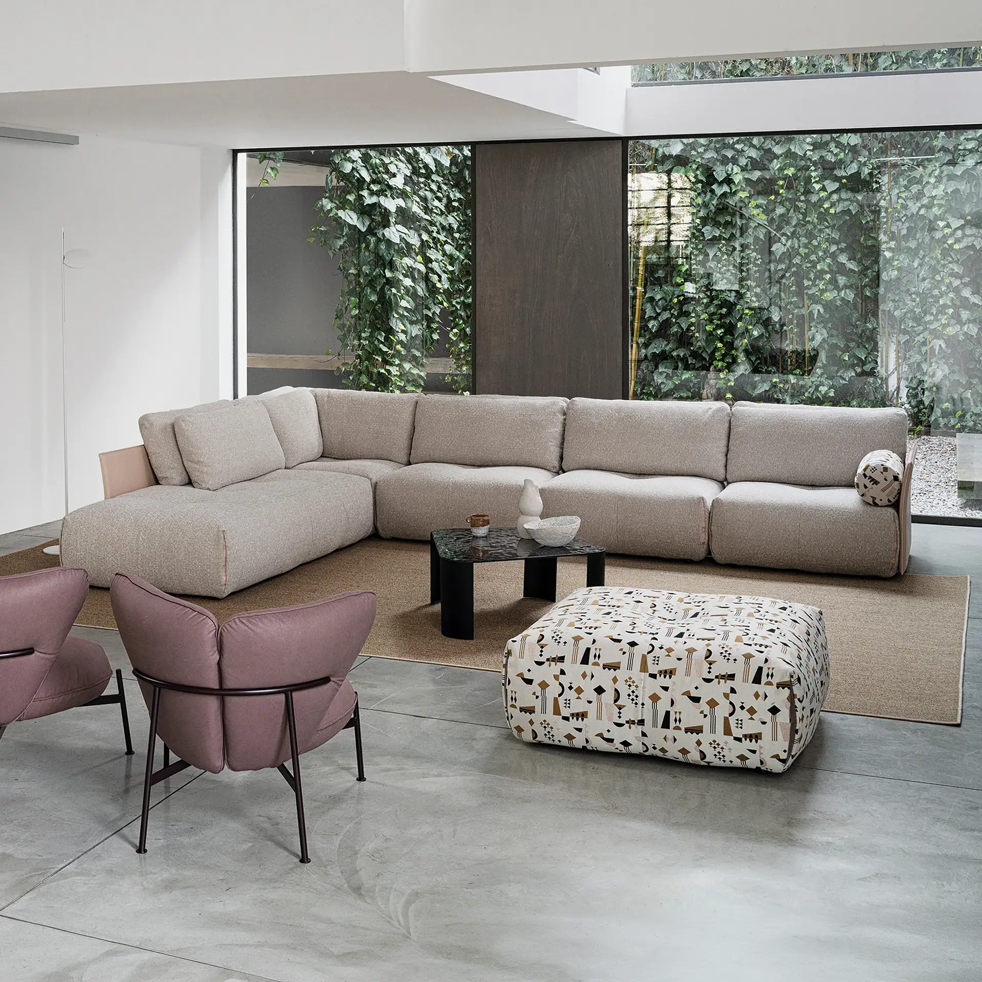 A modern living room with a large beige sectional sofa, two pink armchairs, a patterned ottoman, and a coffee table, all set against a backdrop of floor-to-ceiling windows revealing lush green ivy.