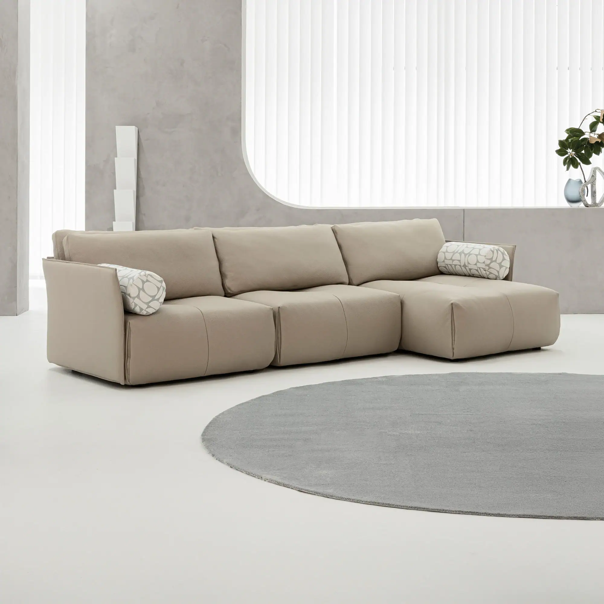 A modern living room with a beige L-shaped sectional sofa, a round gray rug, and a large window with white blinds.