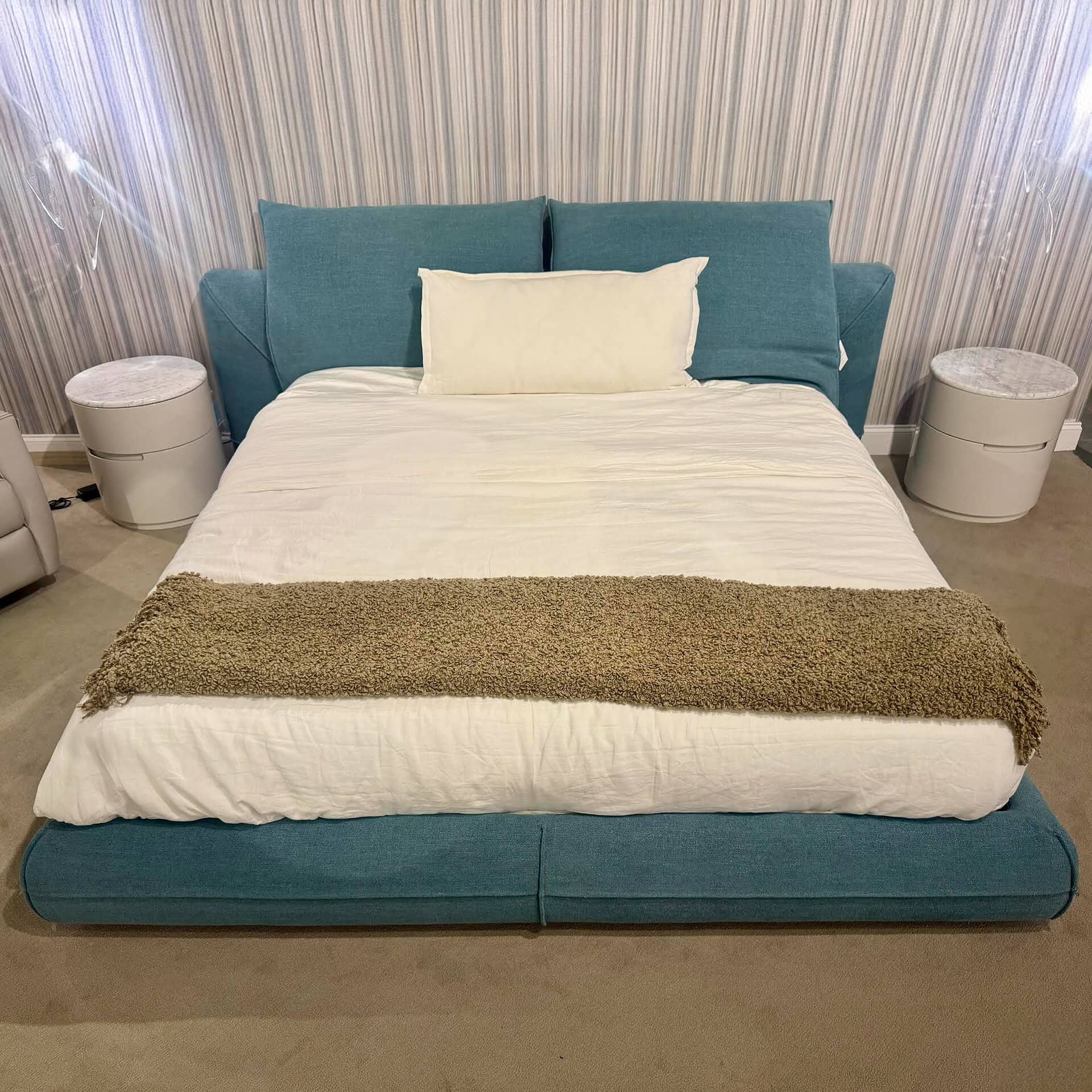 A modern bedroom with a teal upholstered bed, white bedding, and a textured olive-green throw. Two cylindrical nightstands with marble tops flank the bed against a striped wall.