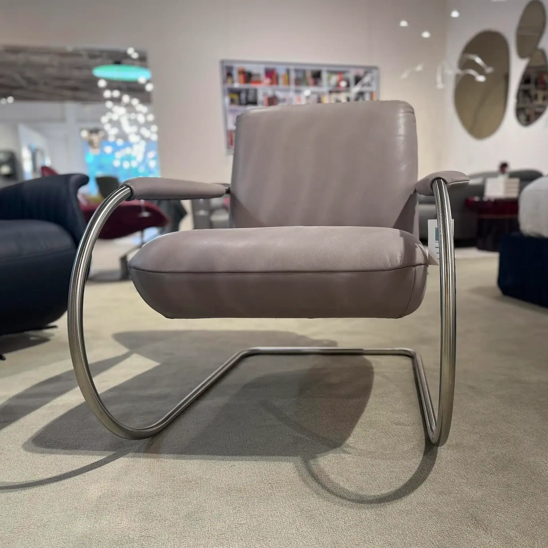 A light gray leather armchair with a polished chrome tubular frame sits on a beige carpet in a furniture showroom.