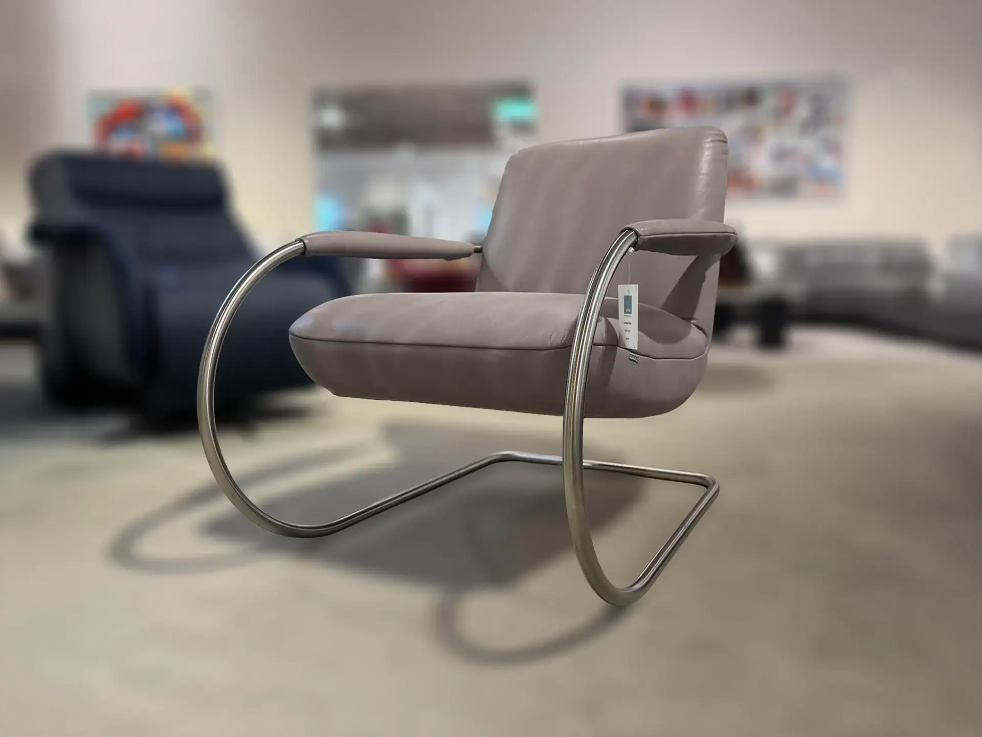 A modern light taupe armchair with a polished chrome cantilever frame in a blurred furniture showroom.