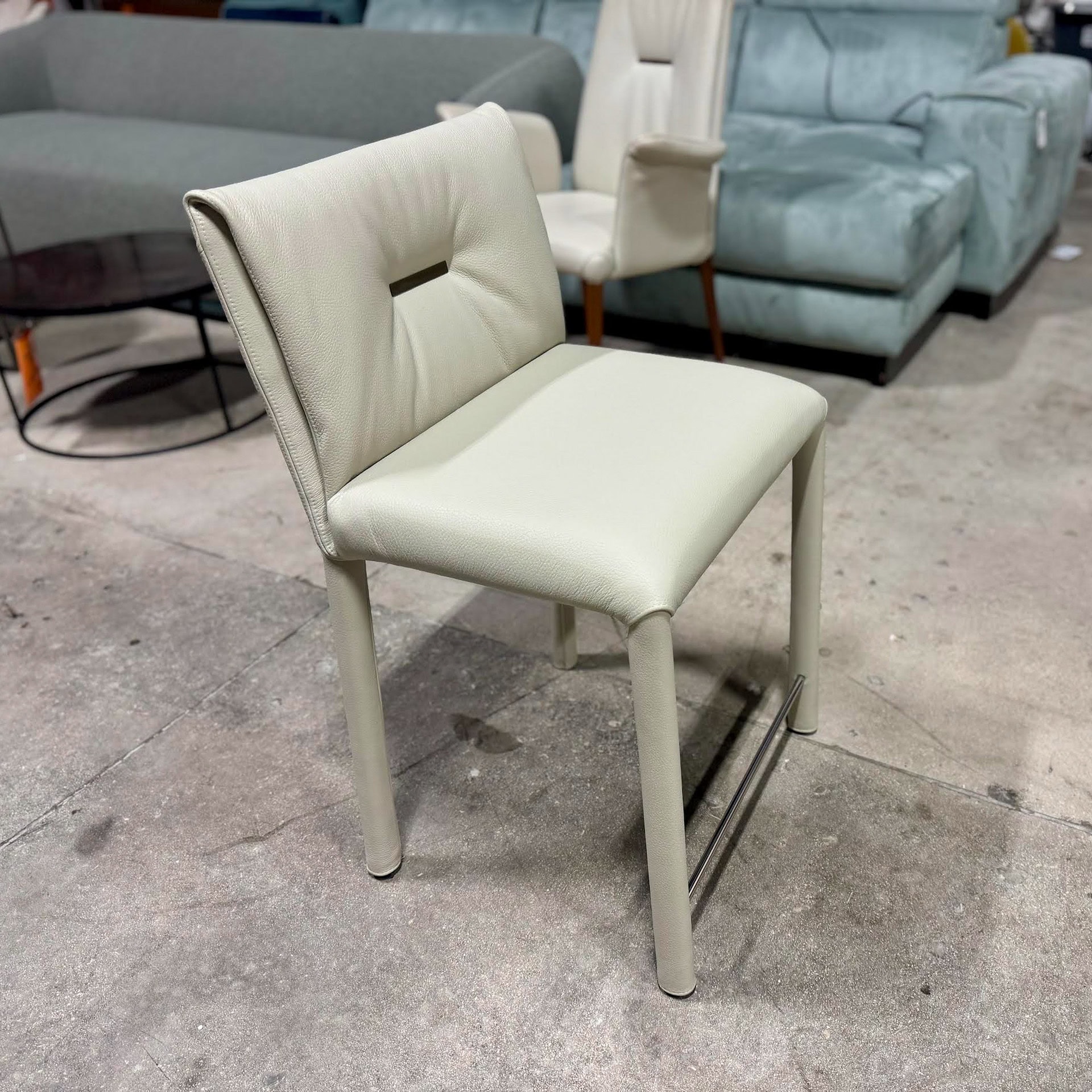 A modern light beige leather counter stool with a cutout backrest and metal footrest, set on a concrete floor in a furniture showroom.