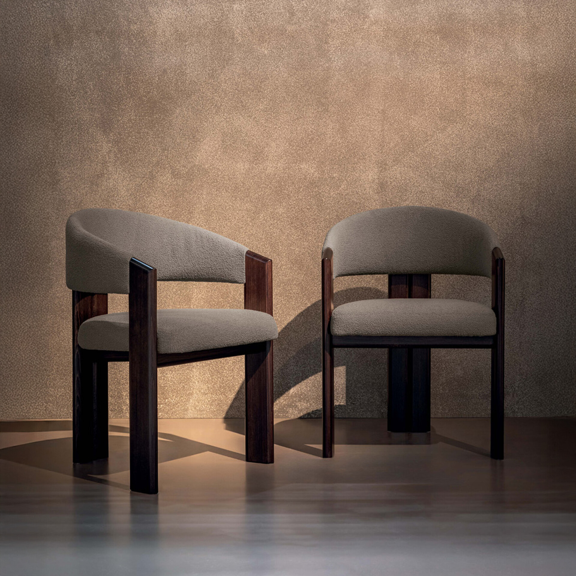 Two modern chairs with dark wood frames and textured grey upholstery, one asymmetrical and one symmetrical, against a textured brown wall with dramatic lighting.