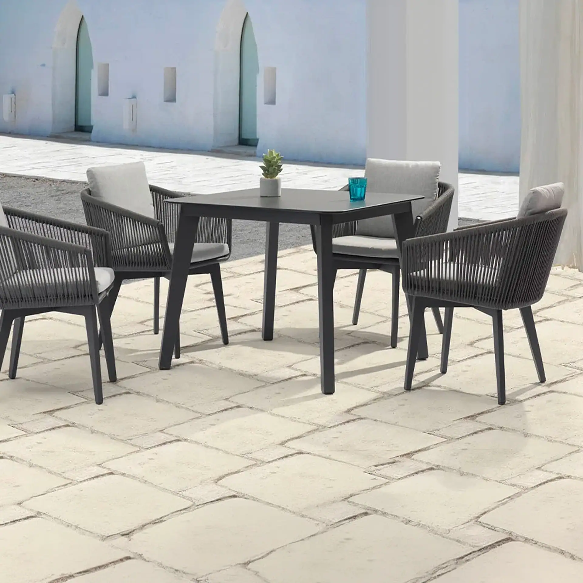 A dark gray square patio table with four matching chairs, each with light gray cushions, on a stone paver patio. A succulent and blue glass are on the table. A white building is in the background.