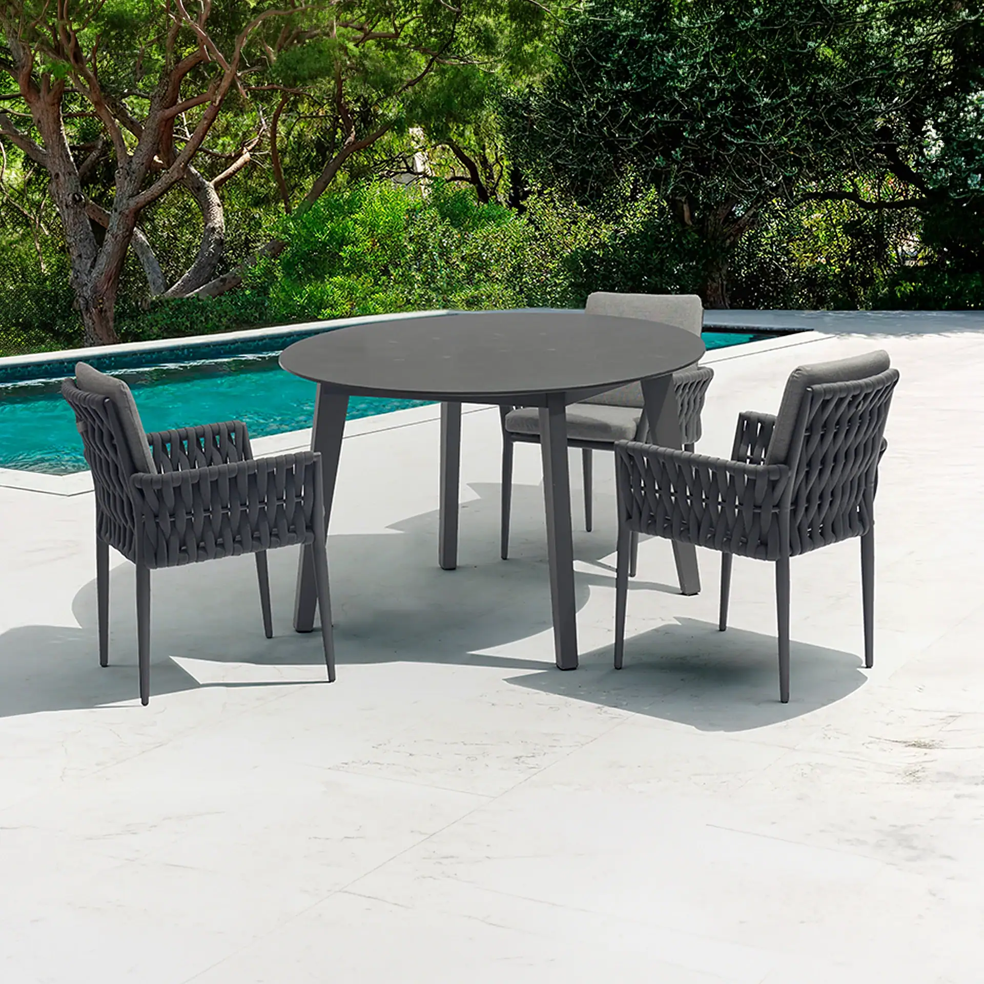 Outdoor dining set with a dark gray oval table and four woven chairs on a light patio next to a turquoise swimming pool, surrounded by green trees.