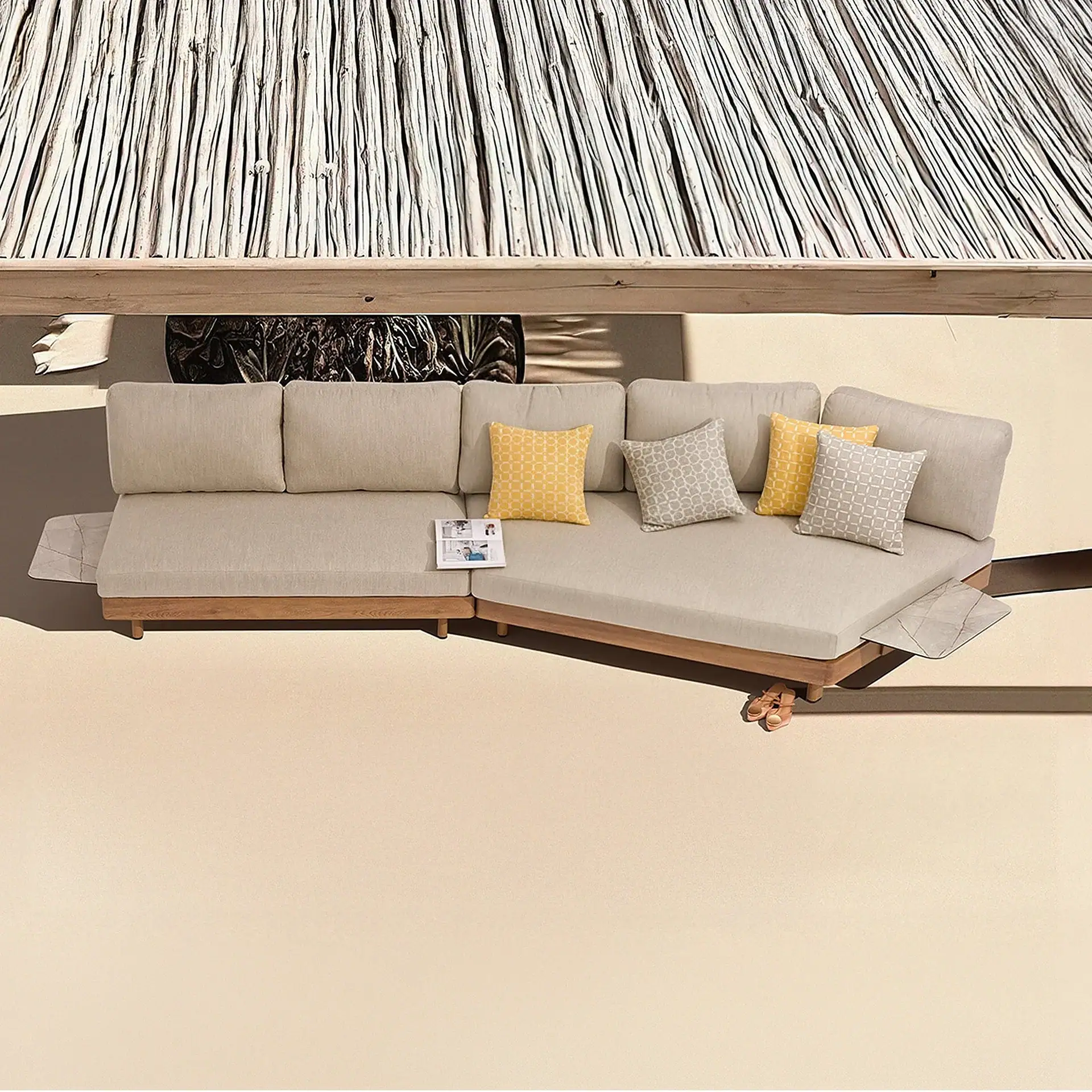 Top-down view of an L-shaped outdoor sofa with light beige cushions, wooden frame, yellow and gray patterned pillows, and a thatched roof overhead on a light patio.