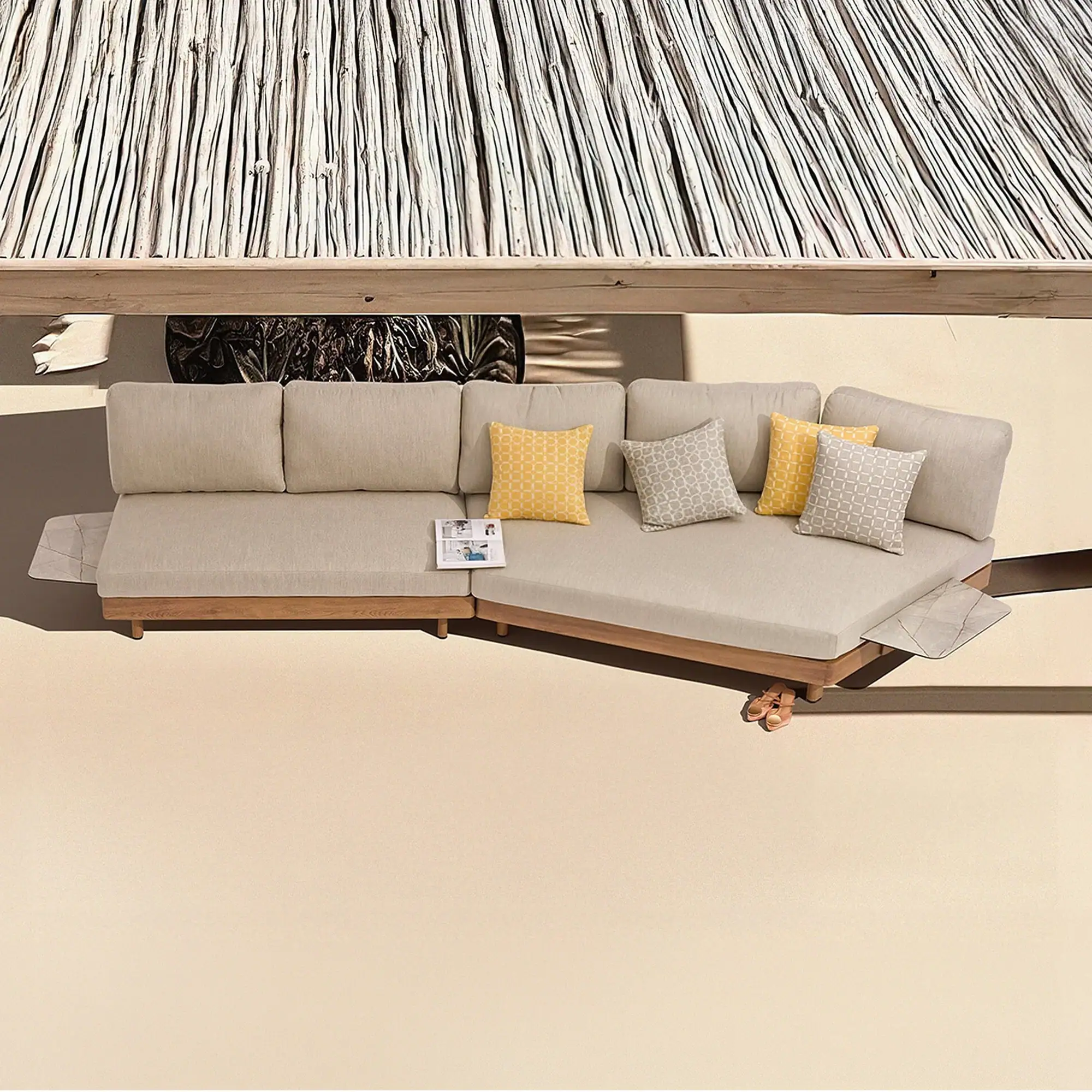 Top-down view of an L-shaped outdoor sofa with light beige cushions, wooden frame, yellow and gray patterned pillows, and a thatched roof overhead on a light patio.