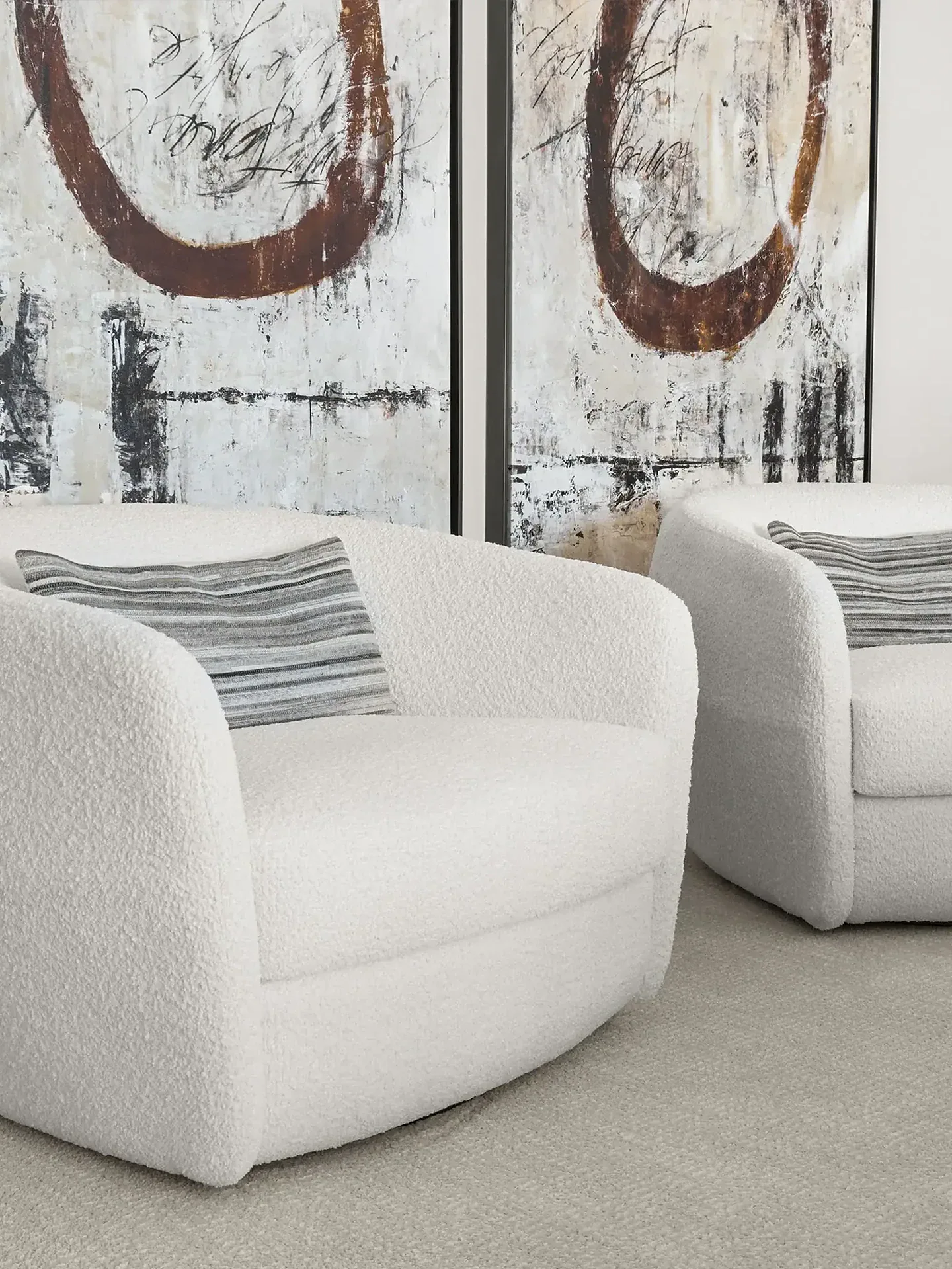 Two white bouclé armchairs with striped pillows on a light gray carpet, in front of abstract artworks.
