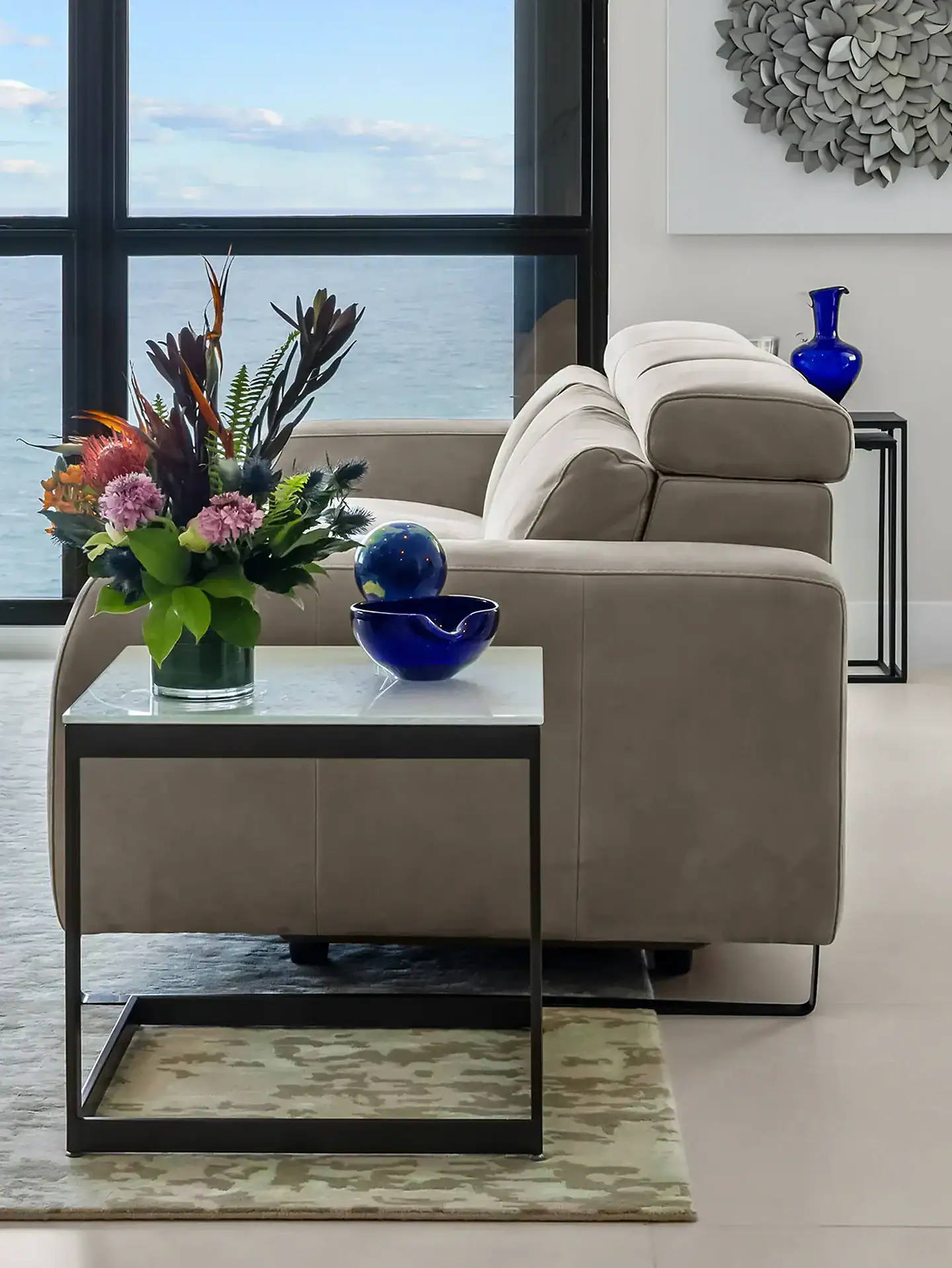 A modern living room with a taupe sofa facing a large window with an ocean view. A side table holds a colorful floral arrangement, a blue bowl, and a blue globe. A grey textured art piece hangs on the wall.