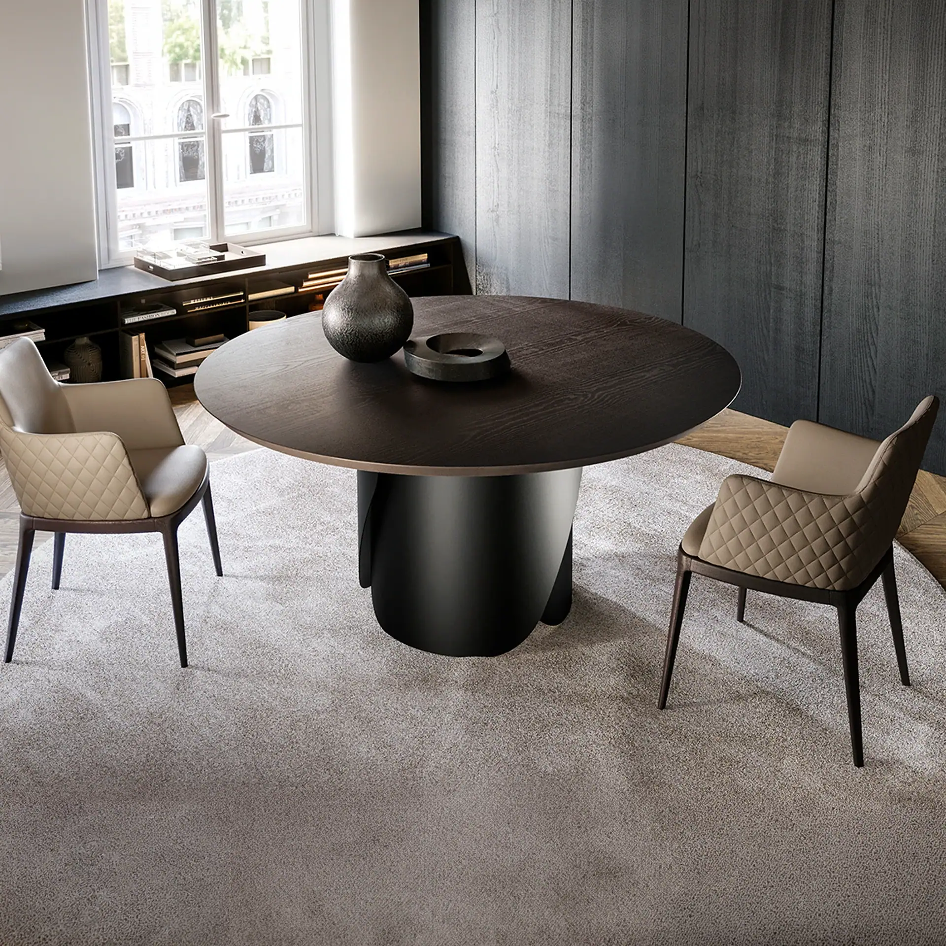 A modern dining room with a round dark wood table, two taupe quilted chairs, a large window, and dark wood paneling.