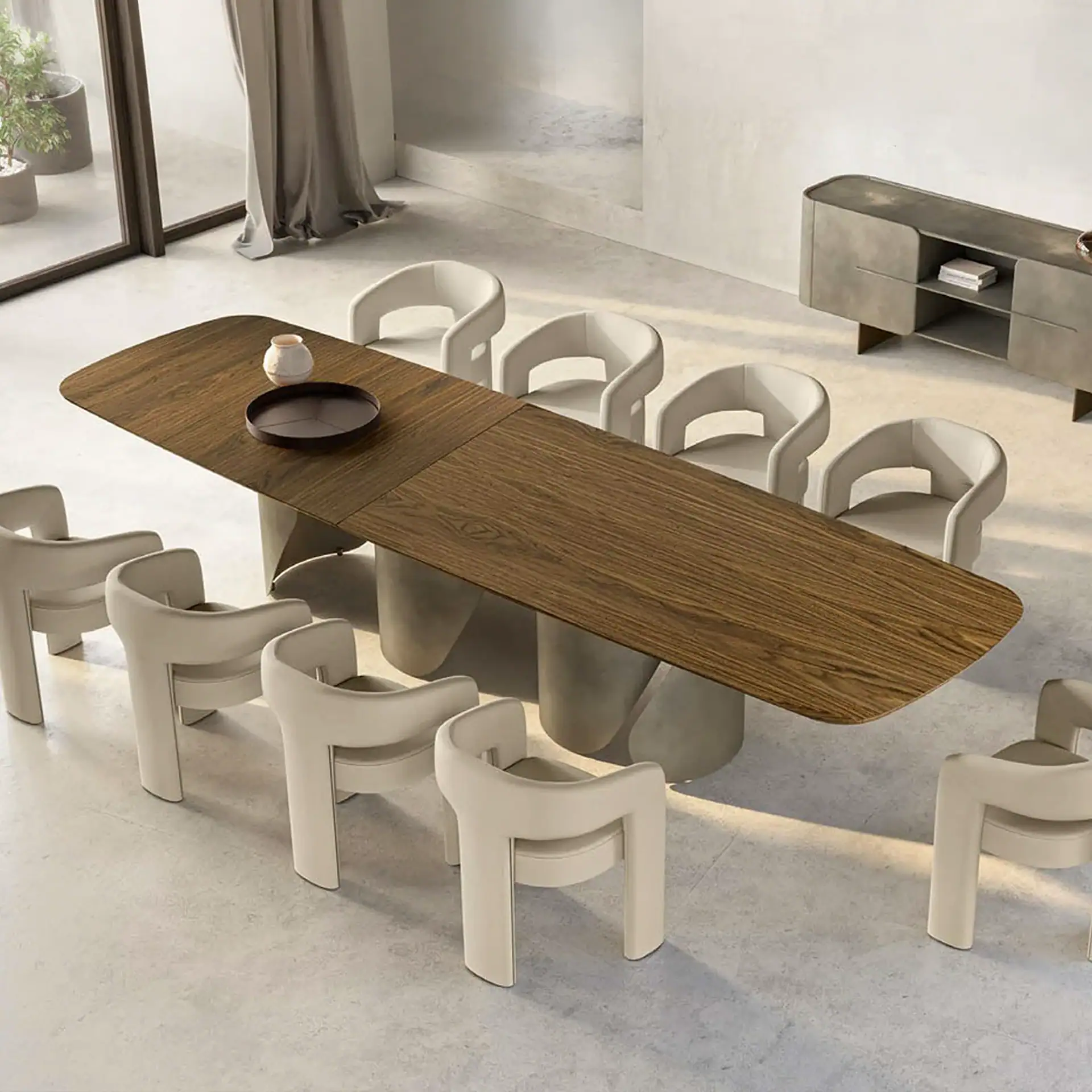 A modern dining room with a long wooden table, ten light beige sculptural chairs, and a grey sideboard on light grey concrete flooring. A large window with a dark frame is visible on the left.