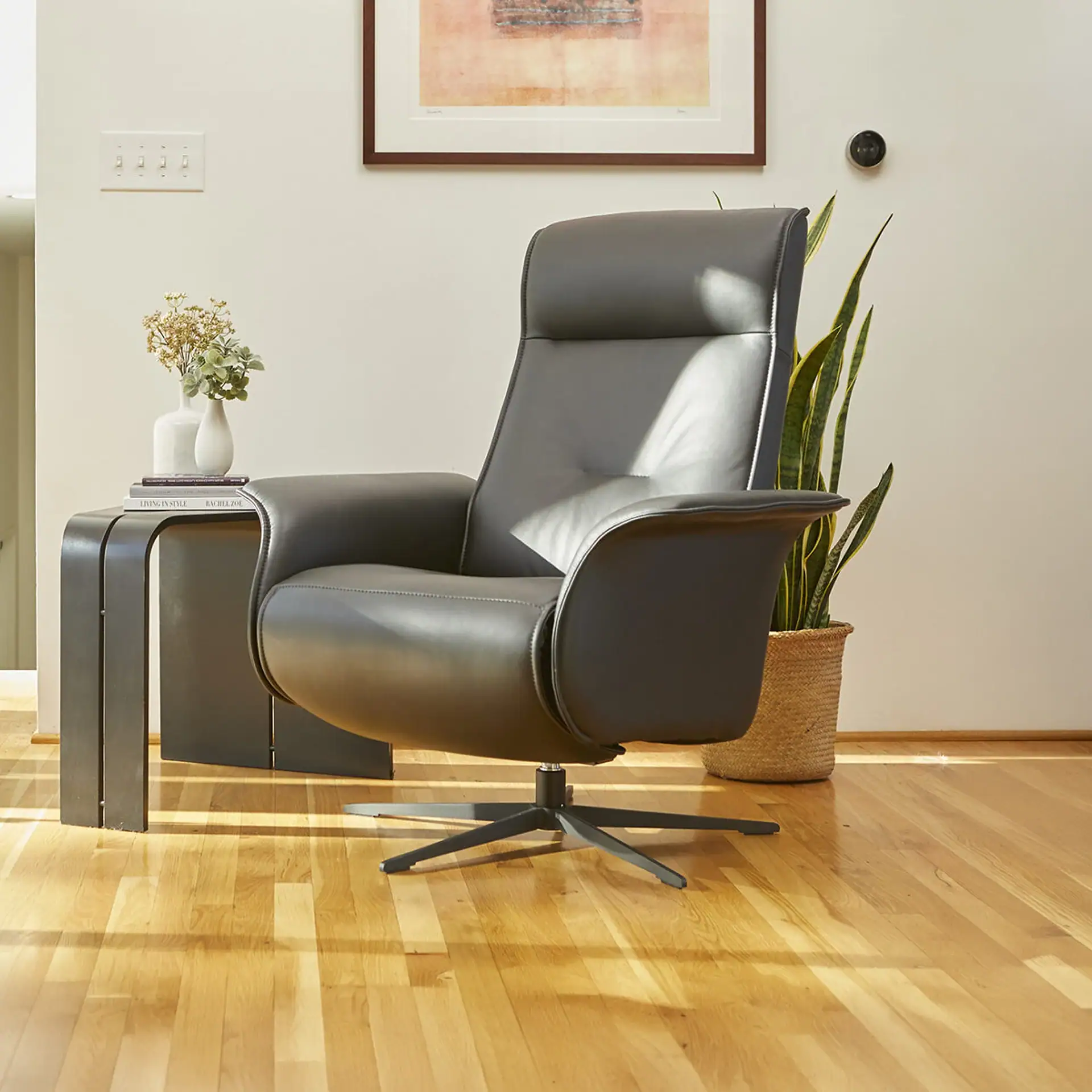 A dark gray leather recliner chair with a swivel base sits on a hardwood floor next to a C-shaped side table with vases and books. A snake plant is on the right, and a framed artwork and smart thermostat are on the wall.