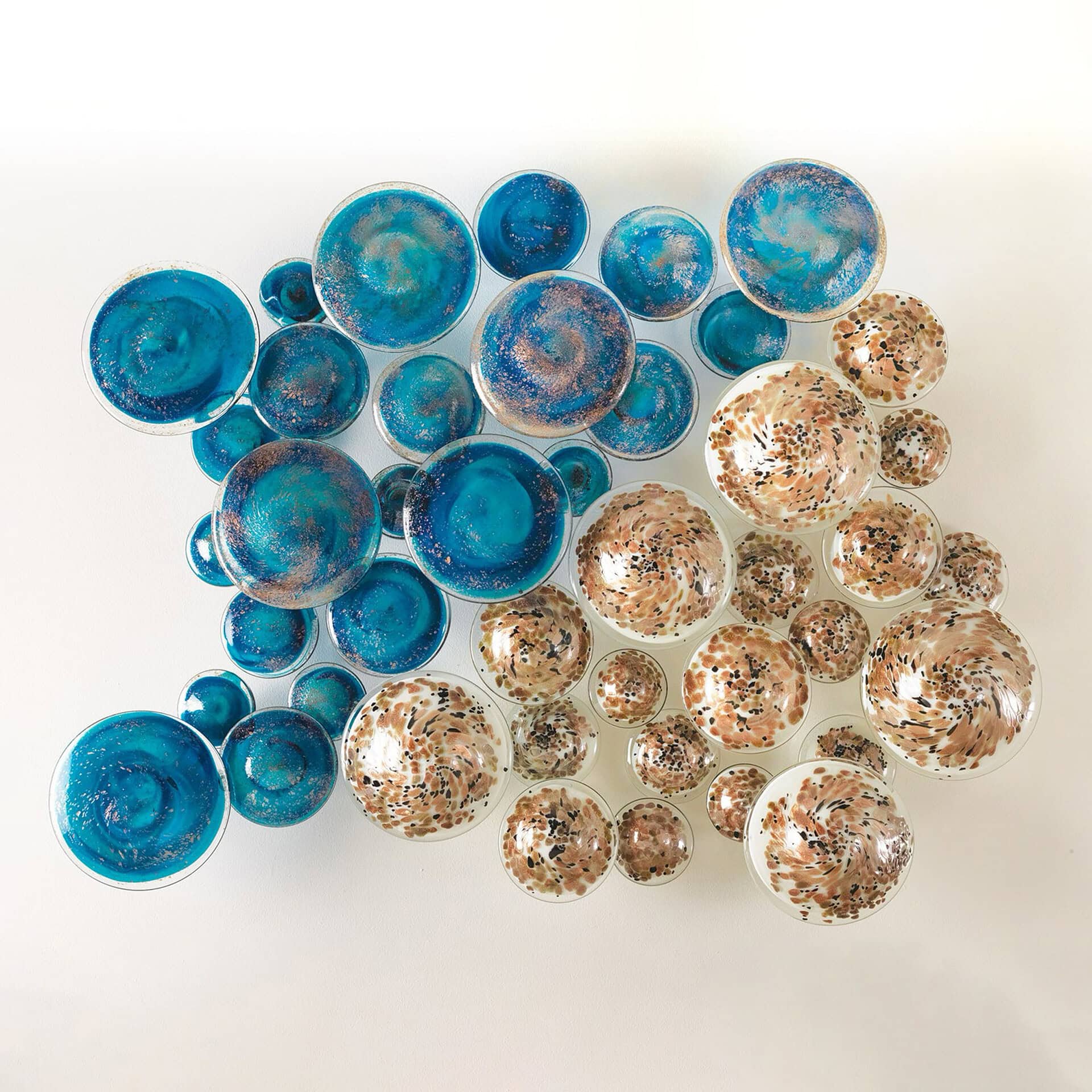 A large wall art installation composed of many circular, dome-shaped glass pieces. The left side features turquoise and teal pieces with metallic flecks, while the right side displays clear glass pieces with brown, copper, and black organic patterns.
