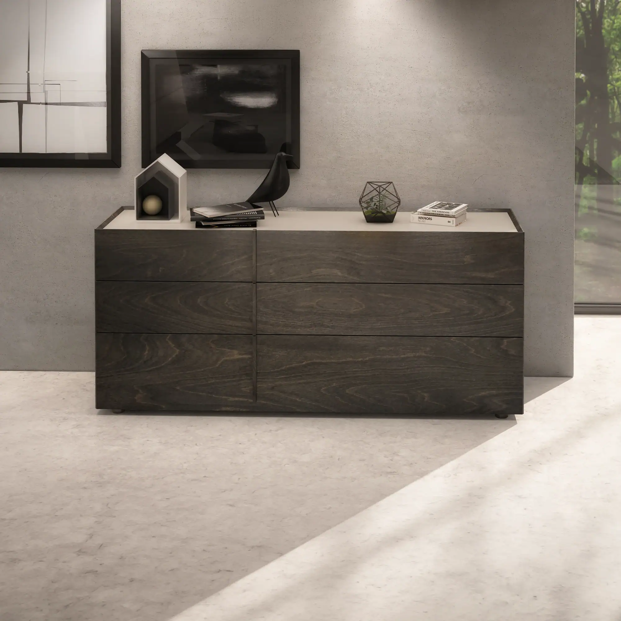 A dark brown wooden dresser with six drawers and a light top, styled with minimalist decor, against a light gray concrete wall with two framed artworks and a window showing trees.