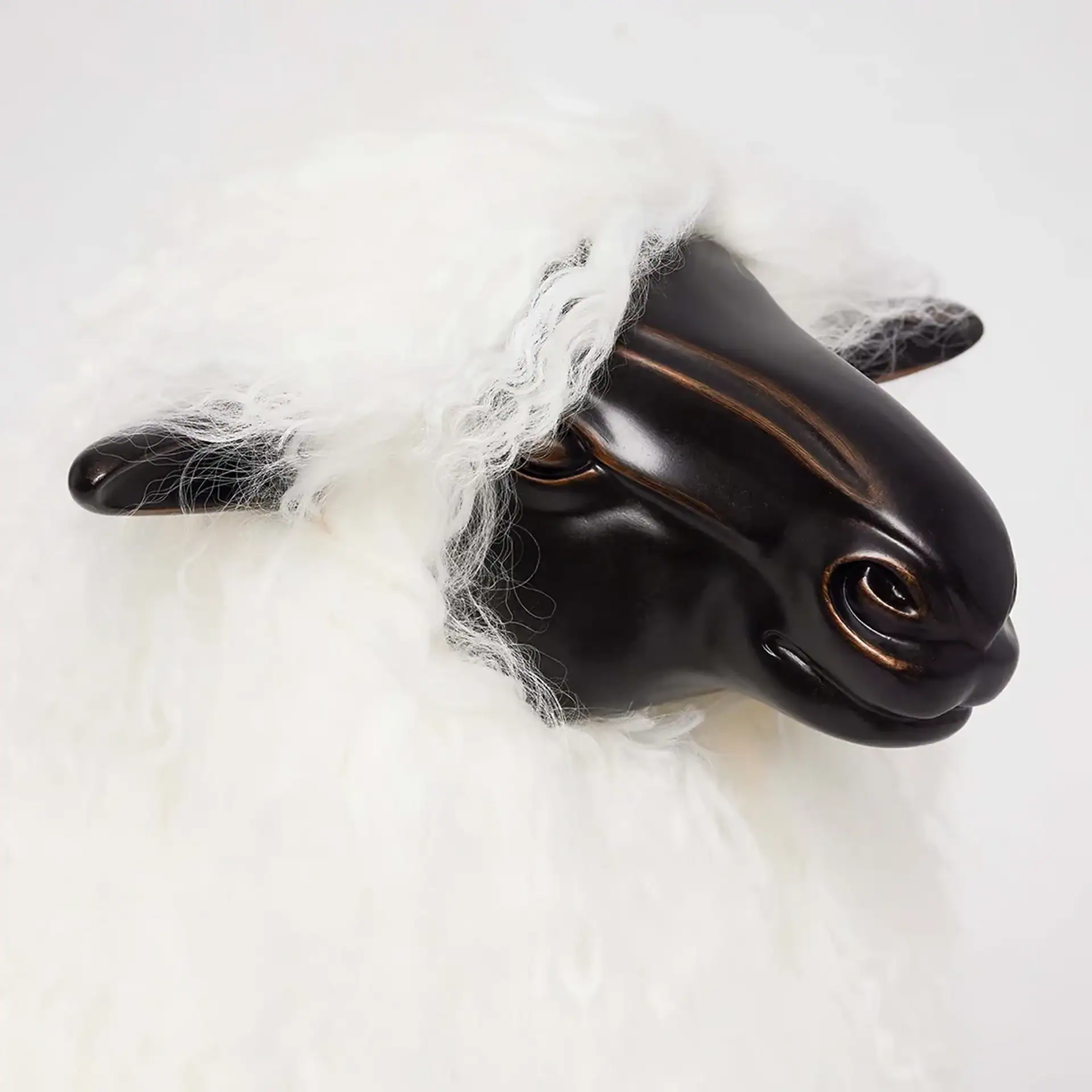 Close-up of a sheep sculpture with a dark, metallic head and fluffy white wool, against a white background.