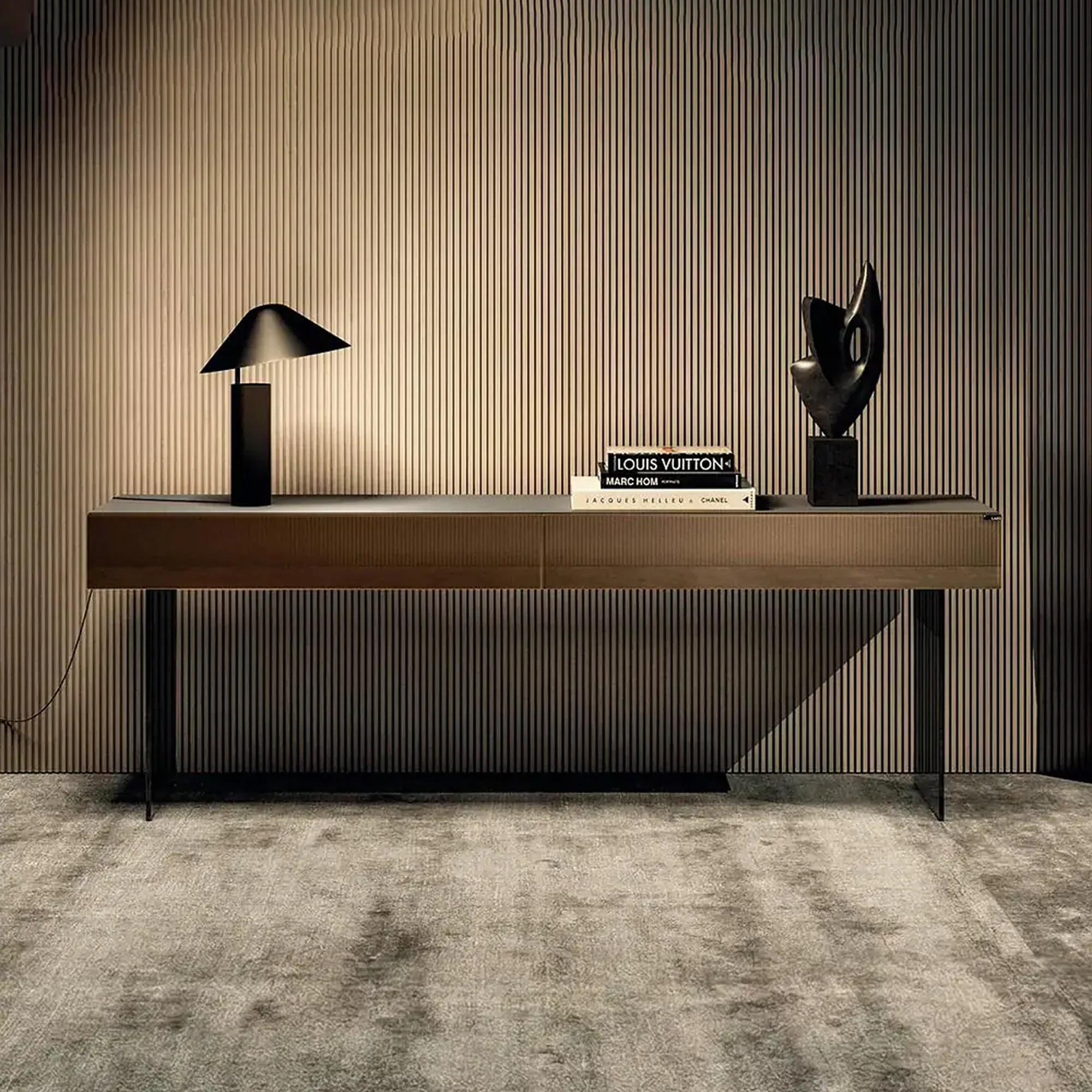 A modern console table with a black lamp, fashion books, and an abstract sculpture against a slatted wall and textured rug.