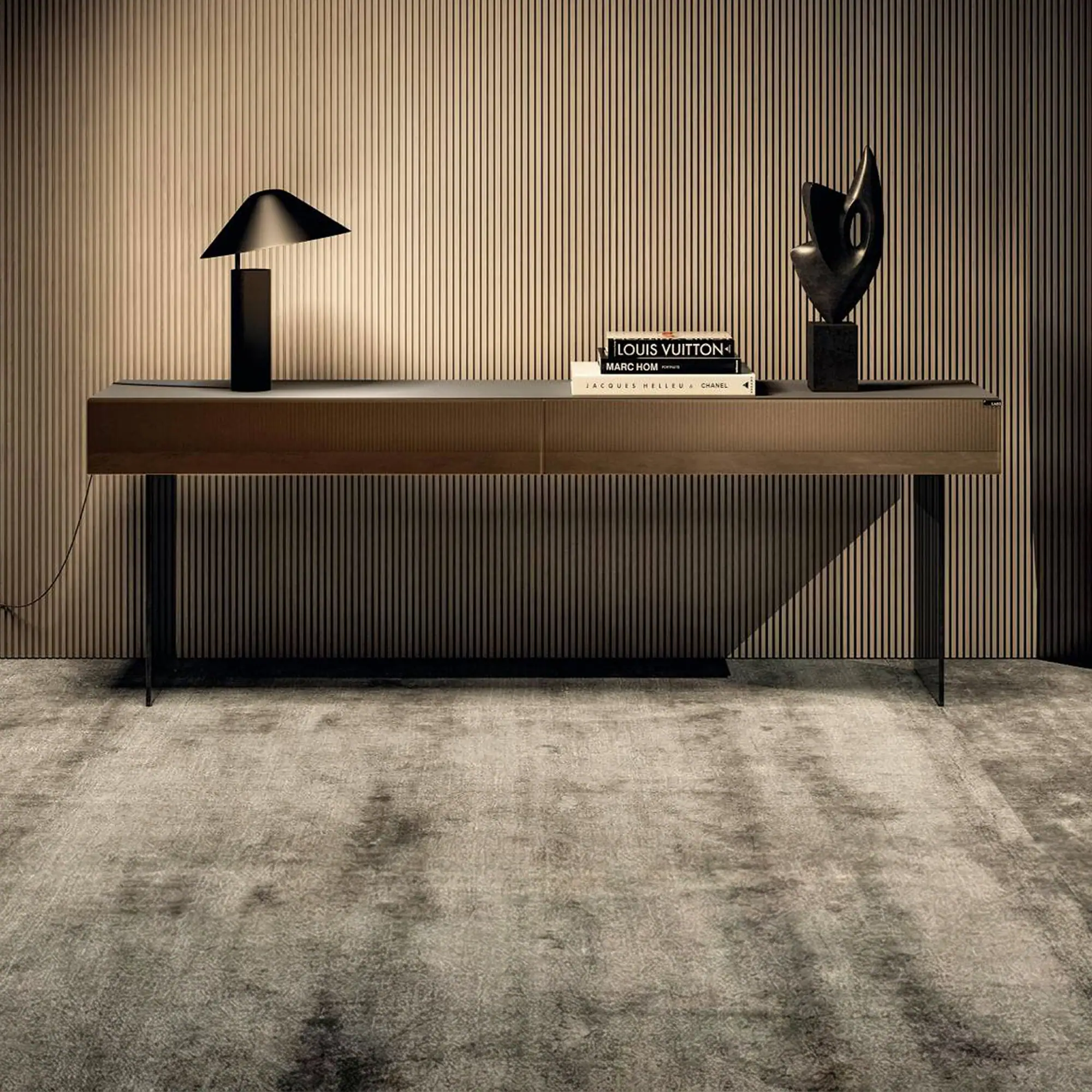 A modern console table with a black lamp, fashion books, and an abstract sculpture against a slatted wall and textured rug.