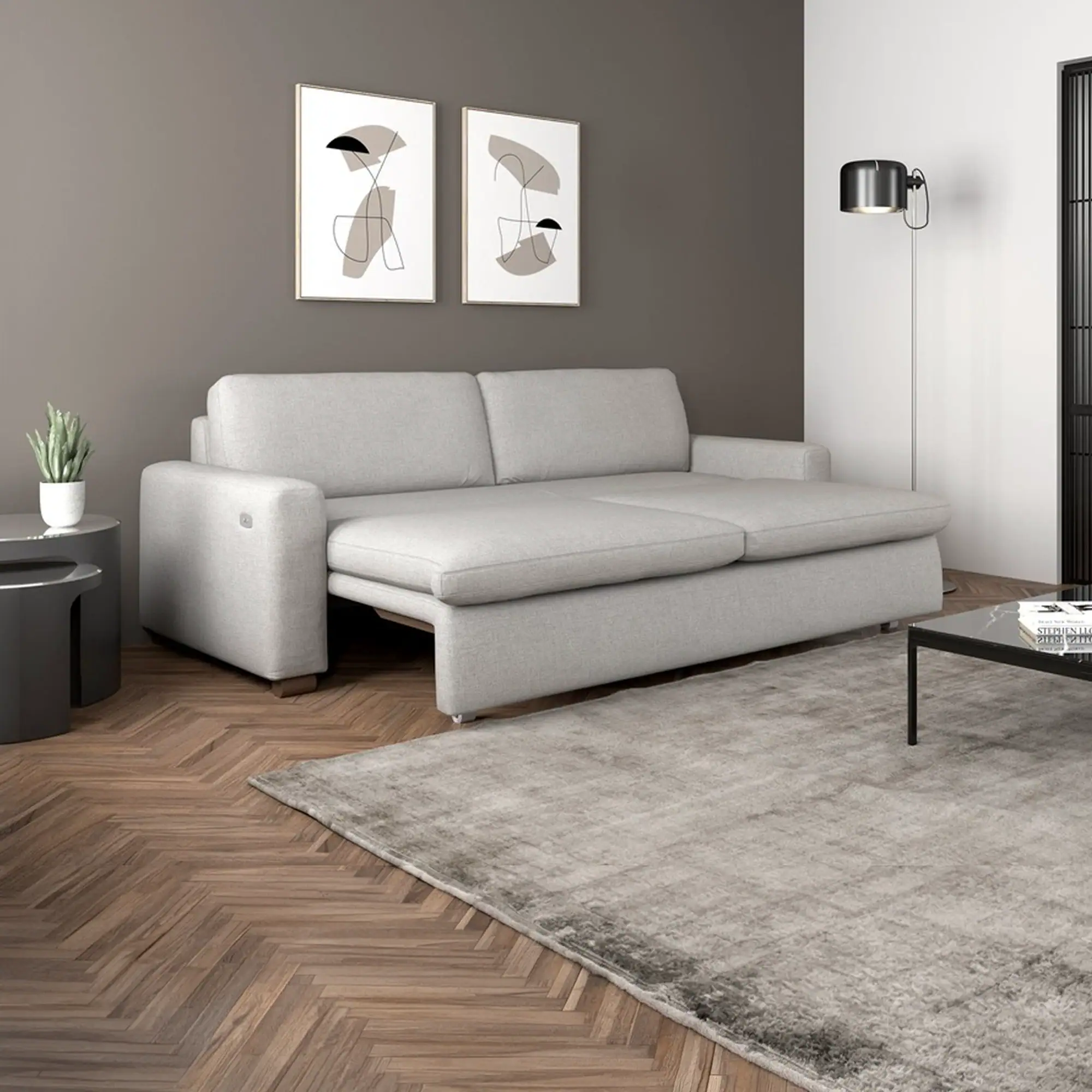 A light gray sofa bed, partially extended, in a modern living room with a dark gray accent wall, abstract art, and a herringbone wooden floor.