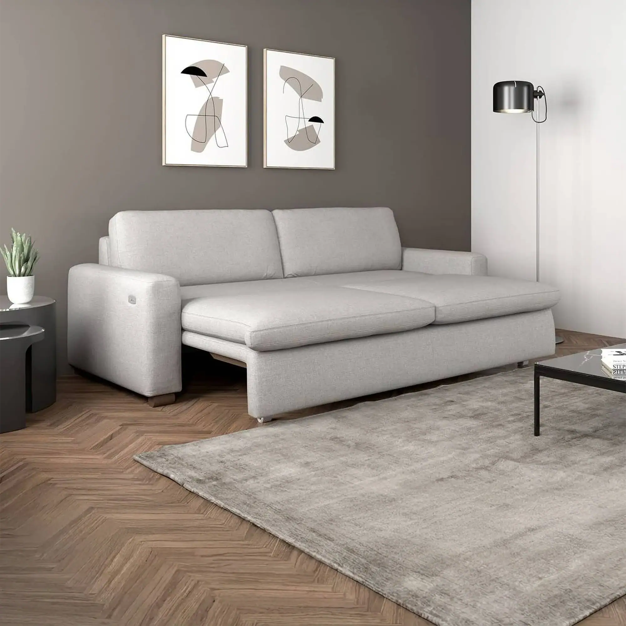 A light gray sofa bed, partially extended, in a modern living room with a dark gray accent wall, abstract art, and a herringbone wooden floor.
