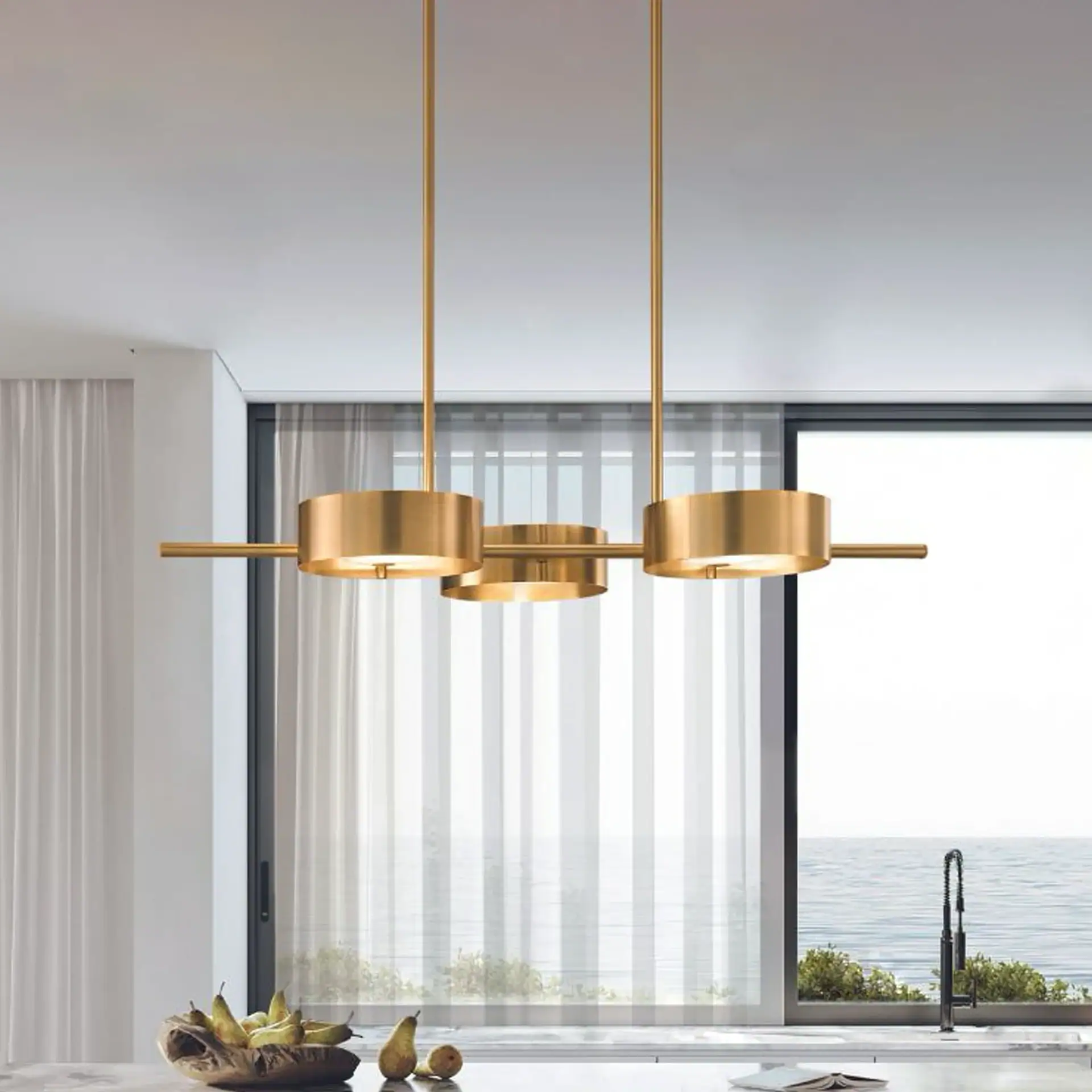 A modern gold chandelier with three cylindrical lights hangs above a countertop with a wooden bowl of pears, set against a large window overlooking a calm blue ocean.
