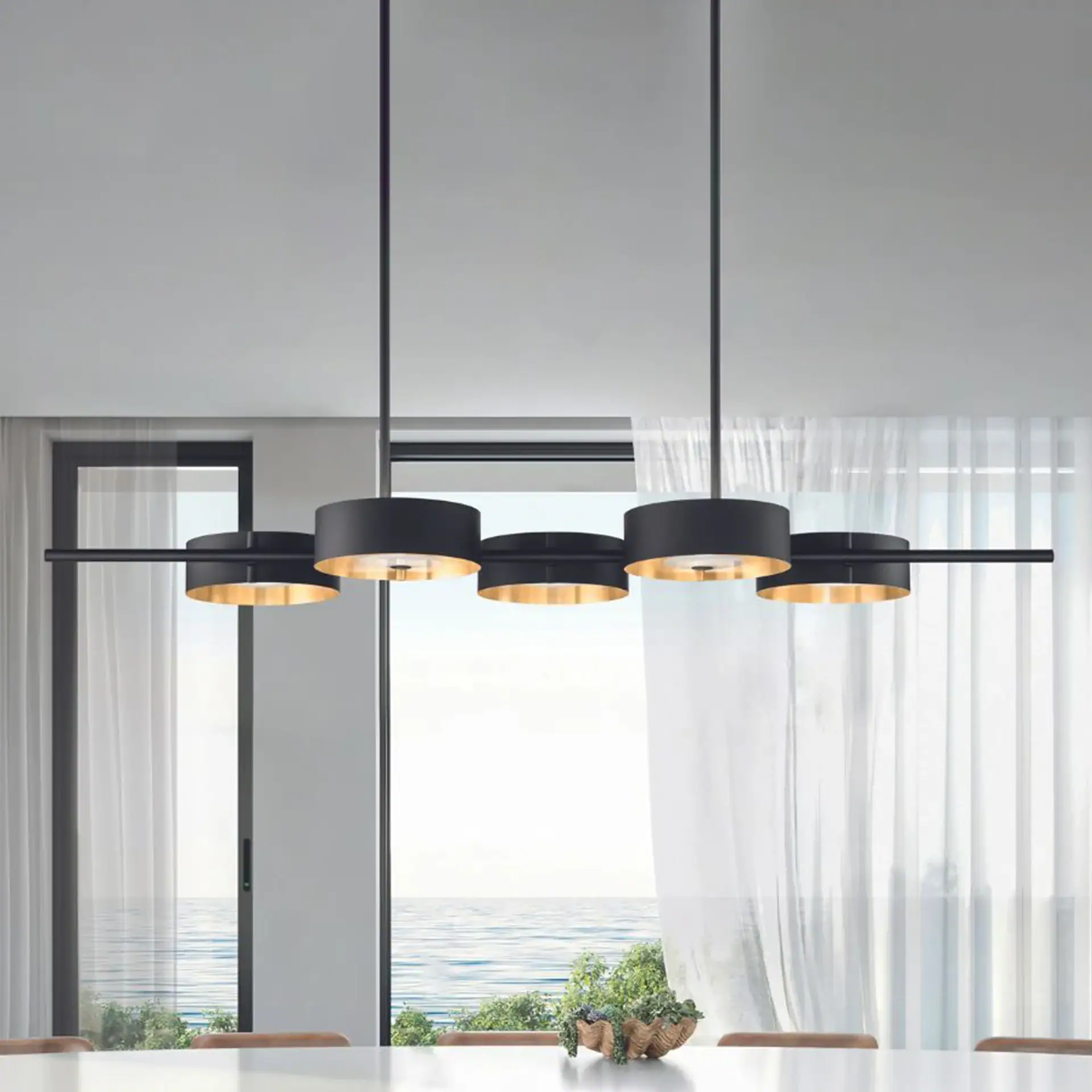A modern black and gold linear chandelier hangs above a white table, with a large window overlooking the ocean in the background.