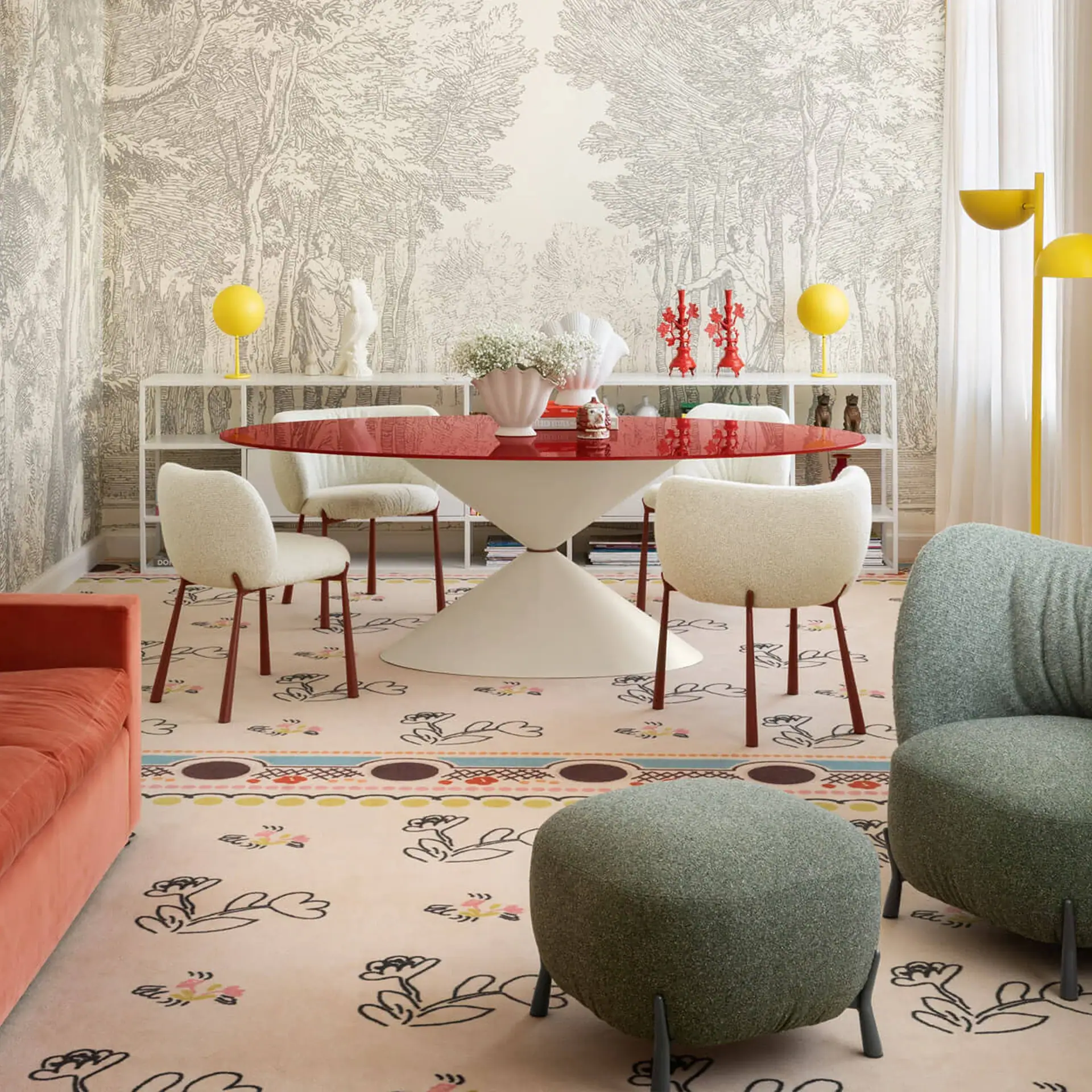 An eclectic room with a red oval dining table, cream chairs, and a pink floral rug. Walls feature grey toile wallpaper. An orange sofa and green armchair are also visible.