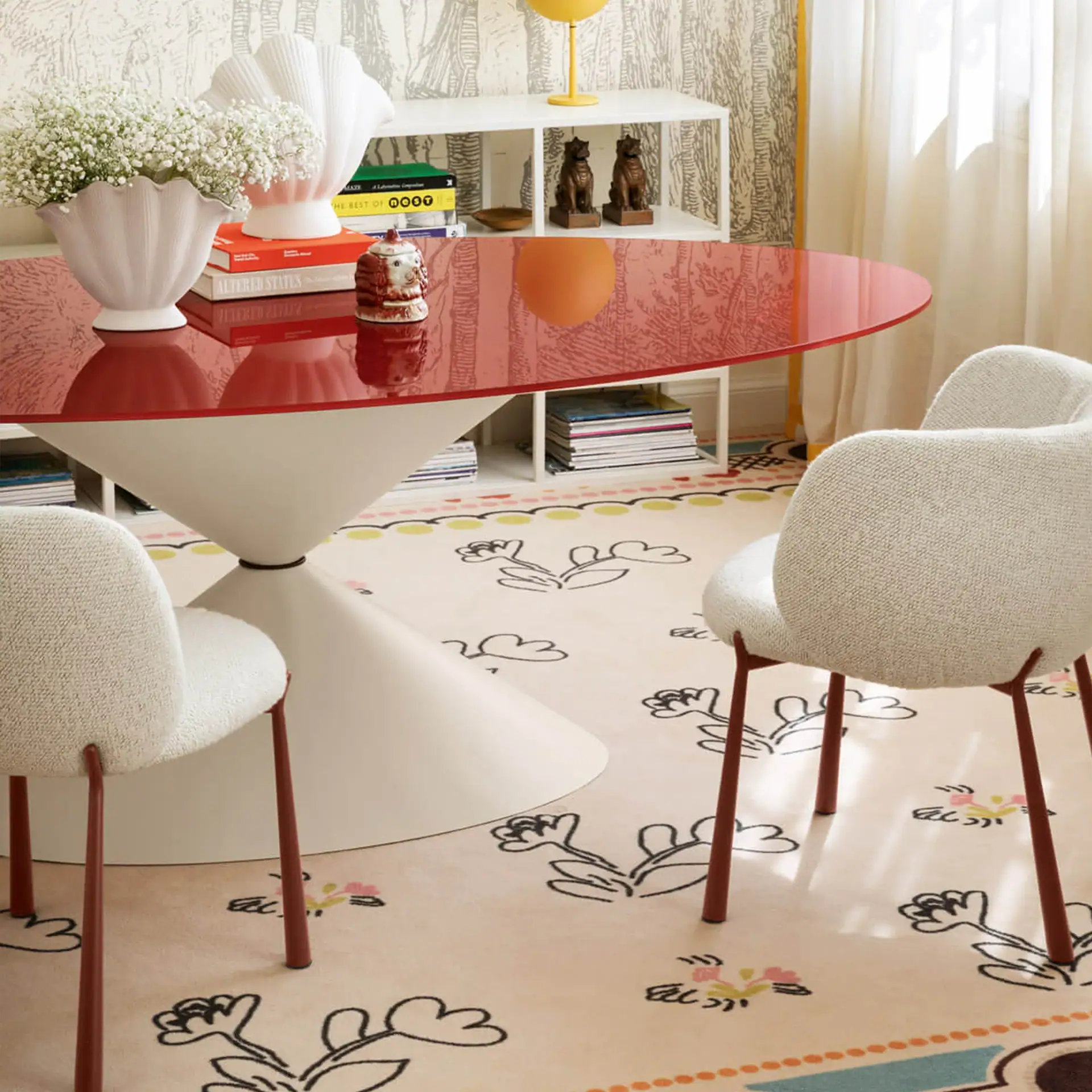 A modern dining room with a red oval table, two cream upholstered chairs, and a floral rug. Decor includes white shell vases with flowers, books, and dog statues.
