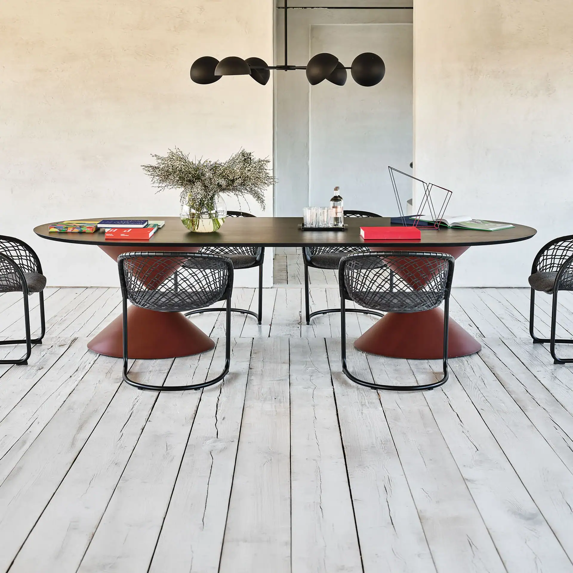 A modern dining room with an oval dark brown table on rust-red bases, surrounded by six black mesh chairs. A black chandelier hangs above. The room has light walls and a distressed white wooden floor.