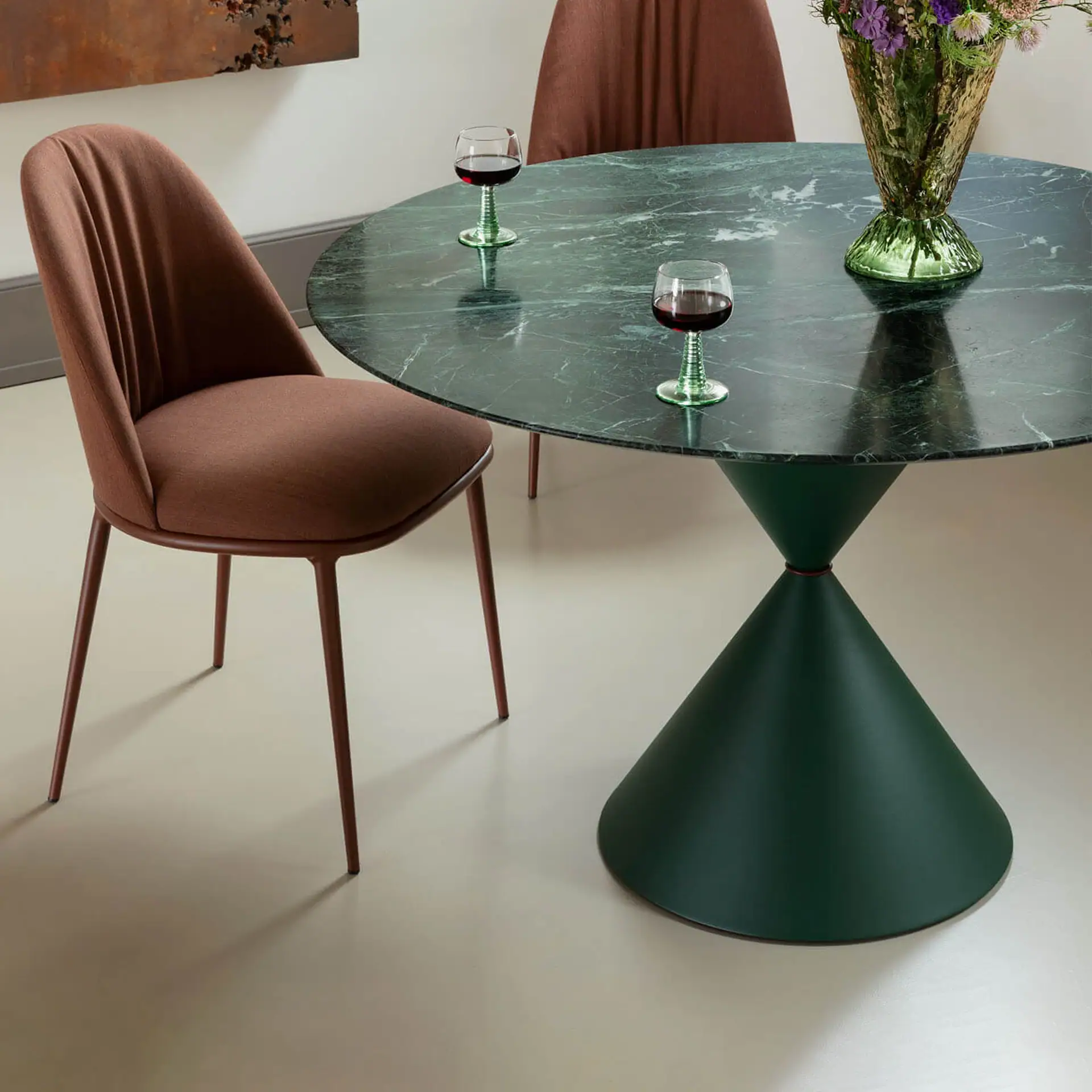 A round green marble dining table with a conical base, two reddish-brown upholstered chairs, two wine glasses, and a vase of flowers.