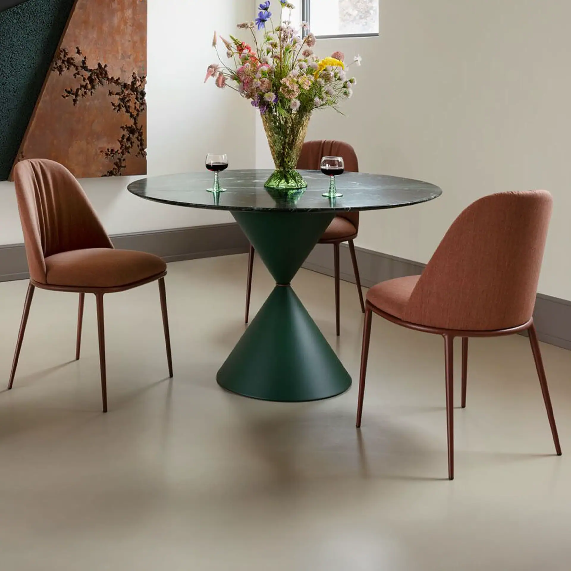 A modern dining table with a dark green marble top and hourglass base, surrounded by three rust-colored chairs. Two wine glasses and a colorful floral arrangement sit on the table. A textured, rusty-orange abstract artwork hangs on the wall.