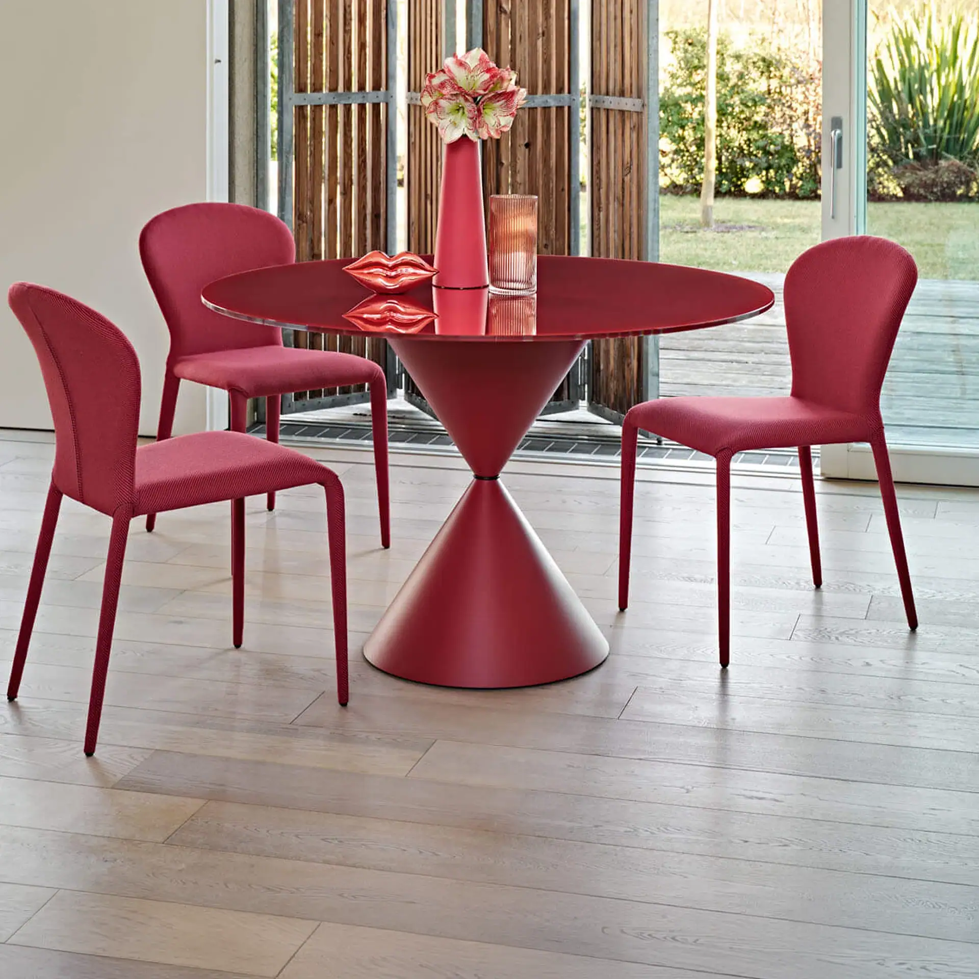 A modern dining room with a red round table and four red chairs on a wooden floor, with a glass door leading to a garden.