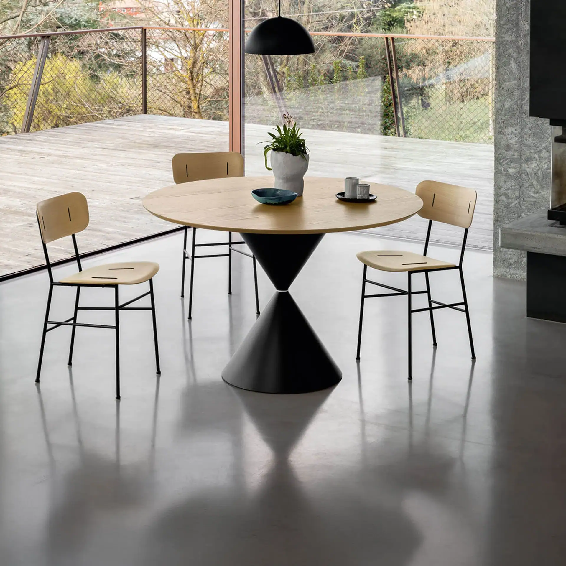 A modern dining room with a round wooden table, black hourglass base, and four light wood chairs on a polished concrete floor, overlooking an outdoor deck and trees through large windows.