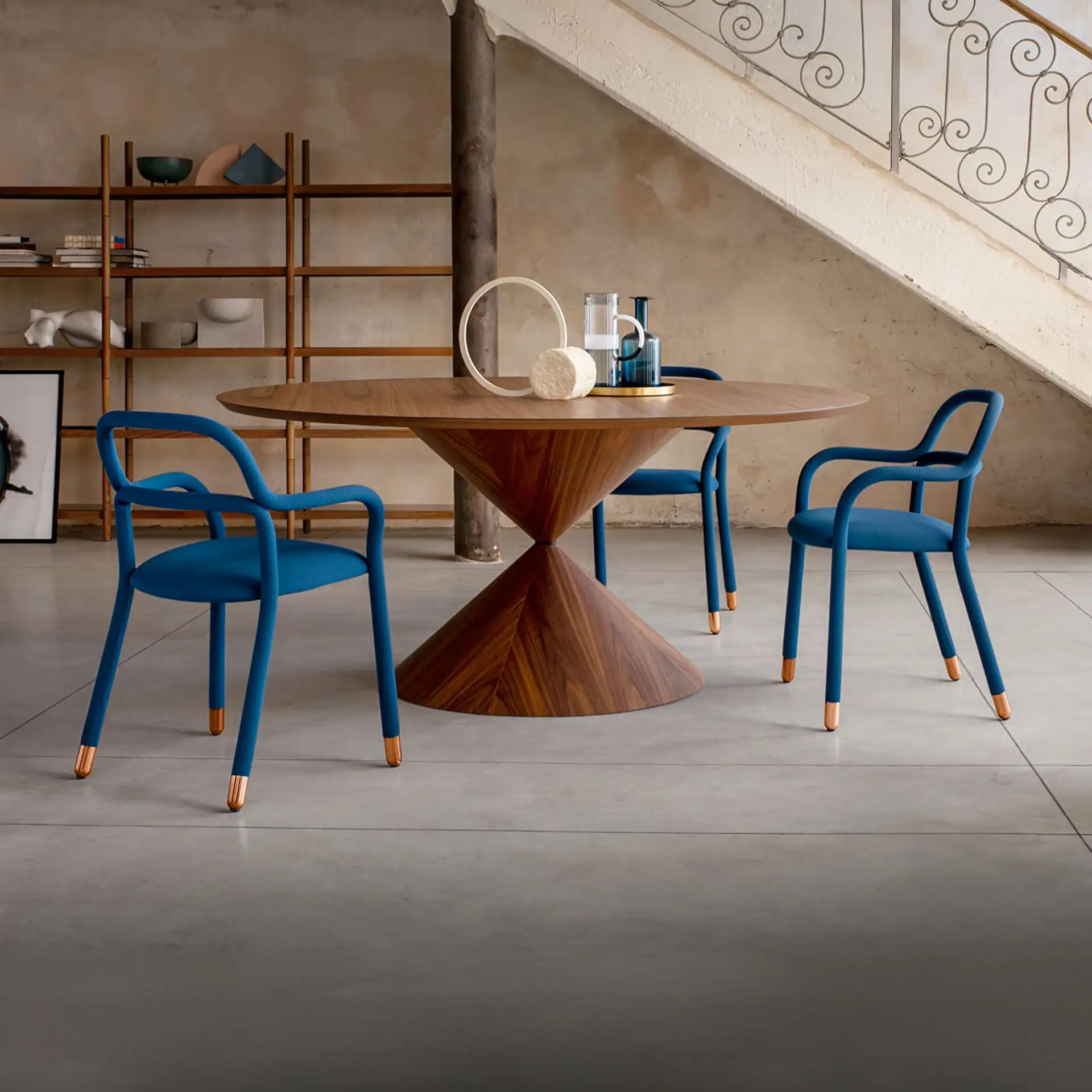 A modern dining room with a round wooden table, three blue upholstered chairs with copper-capped legs, and decorative items on the table. A wooden shelf and a staircase are in the background.