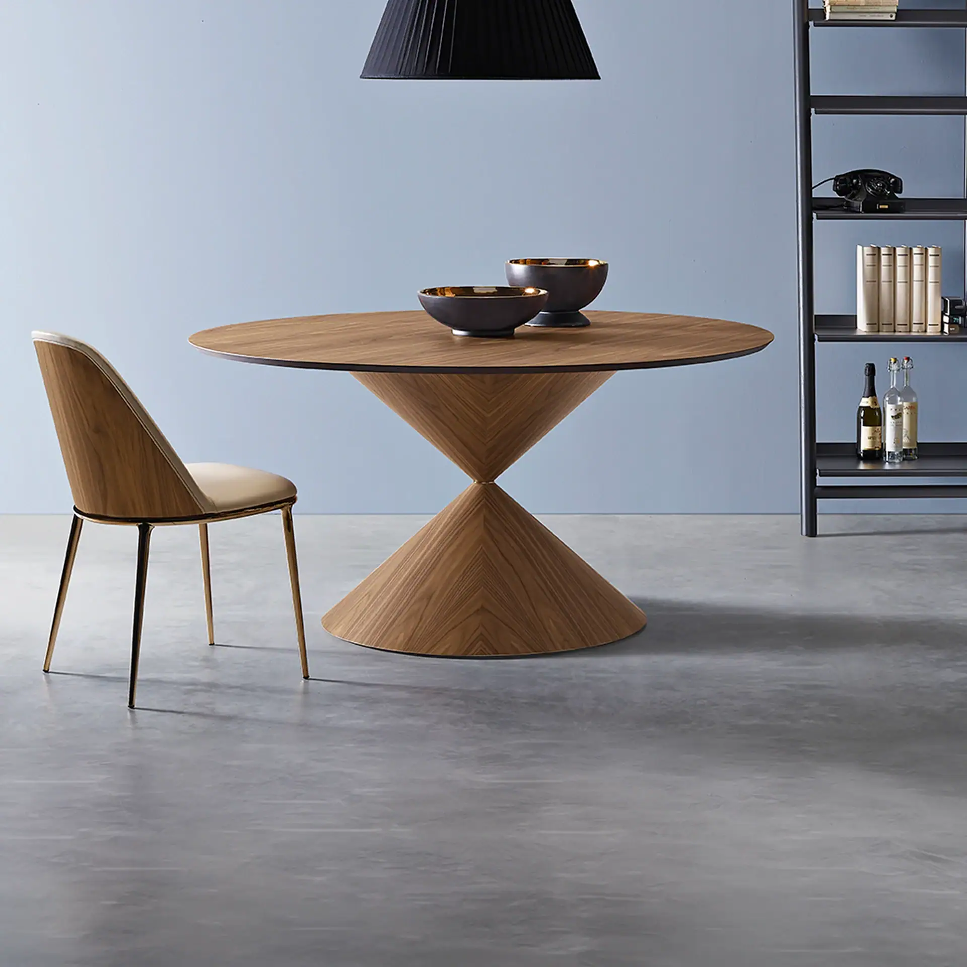 A modern dining table with an hourglass-shaped wooden base and a round wooden top, accompanied by a chair with wooden back and cream seat. A black pendant lamp hangs above, and a black shelving unit with books and a rotary phone is in the background.
