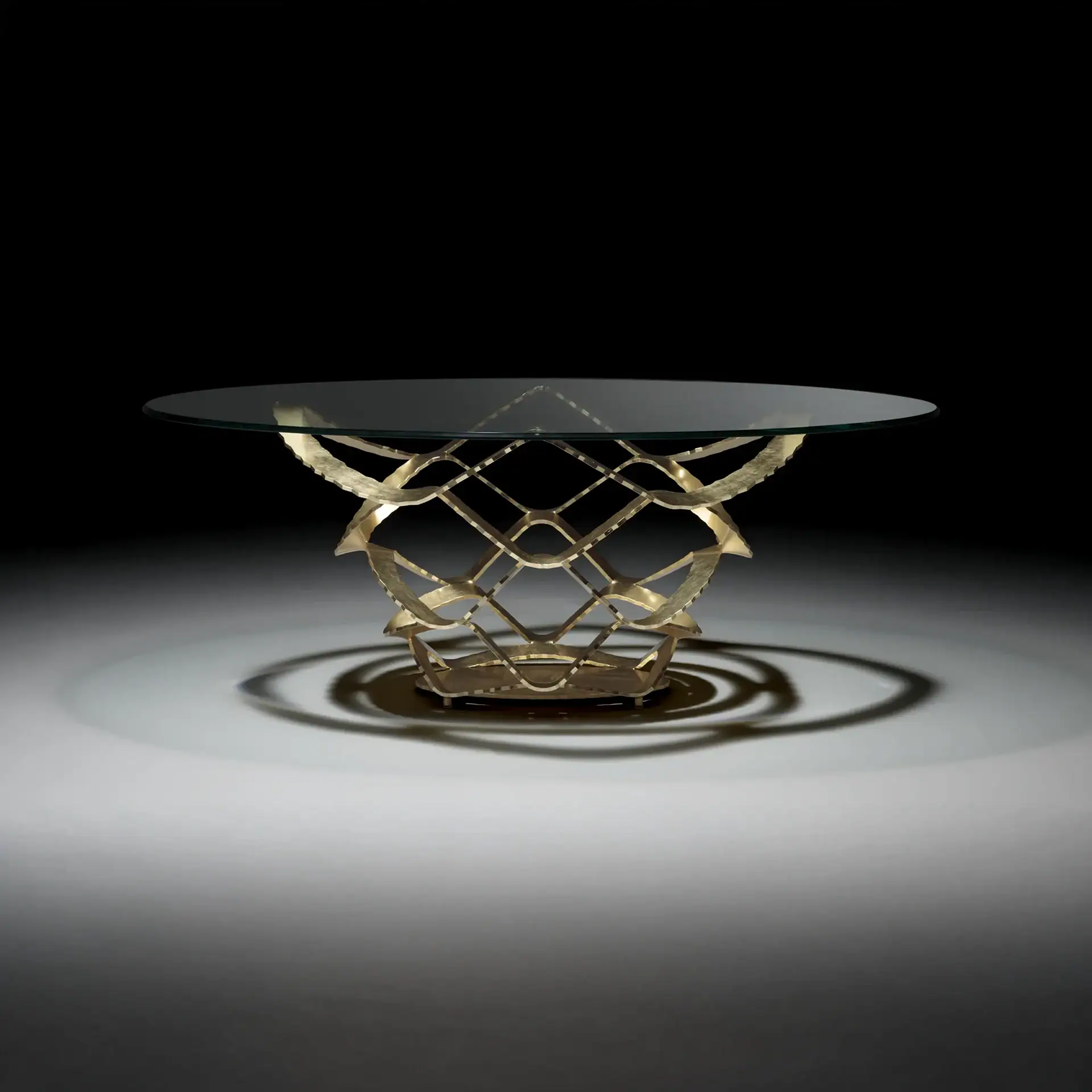A modern coffee table with a clear round glass top and an ornate, textured golden metal base, casting shadows on a light gray surface.