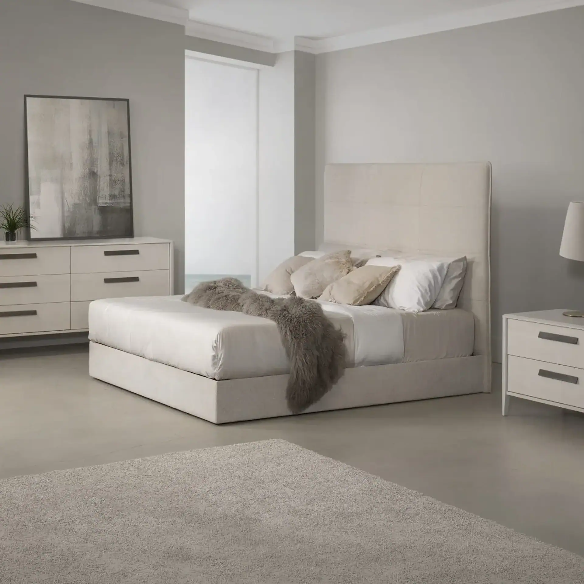A modern bedroom with a cream upholstered bed, white dresser, and nightstand, set against light grey walls and floor with a shag rug.