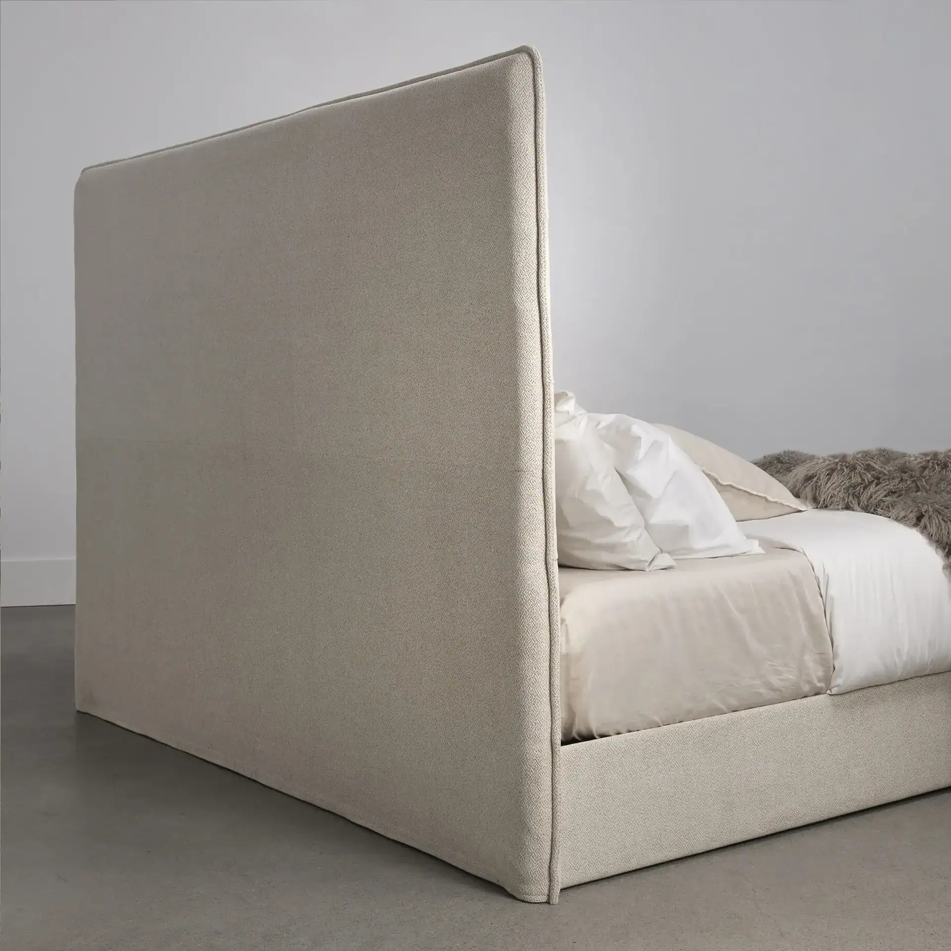 Side view of an upholstered bed featuring a tall, light beige headboard, neutral bedding, and a gray textured throw blanket.